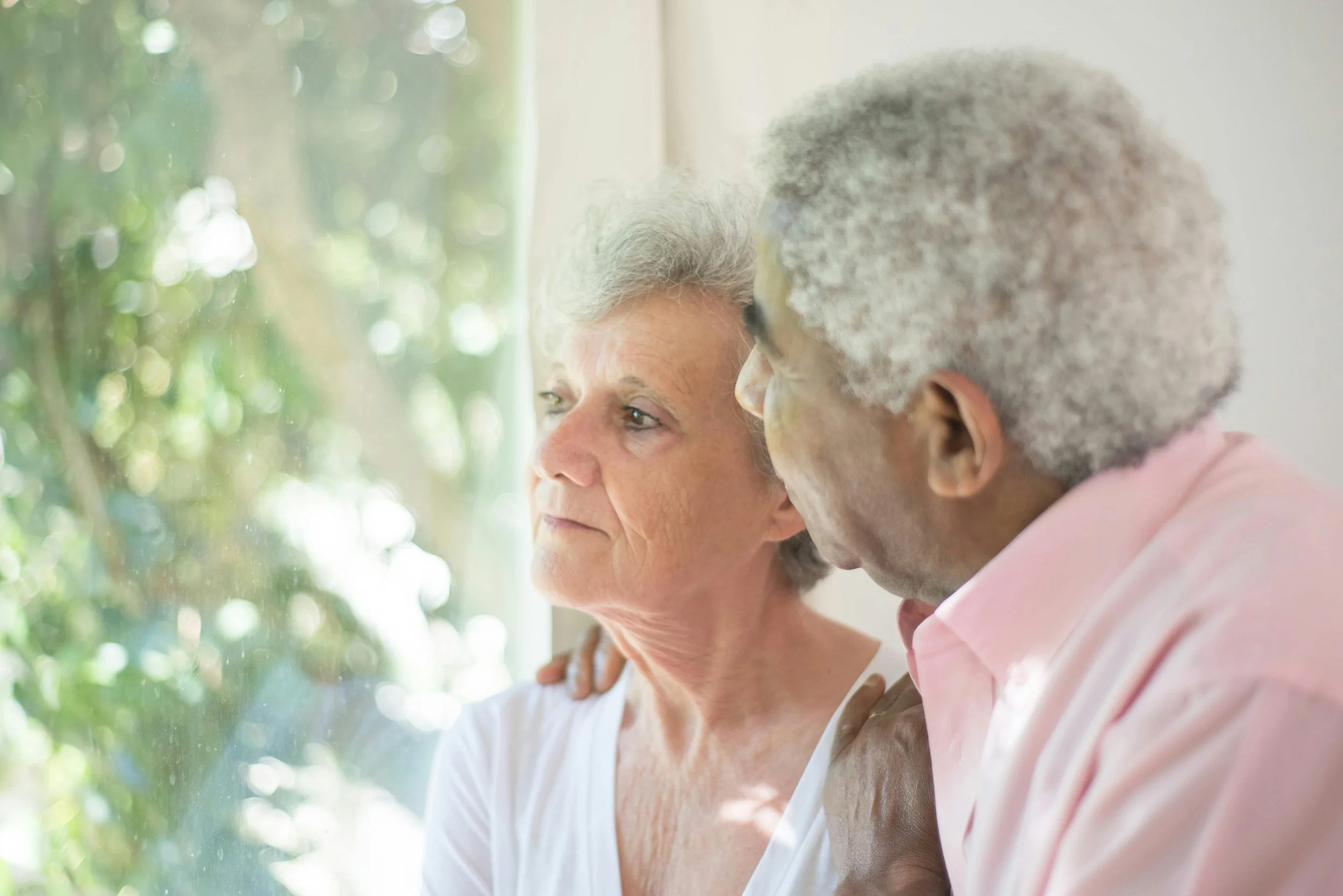 An elderly couple sharing a tender moment indoors by a window with natural light, embracing each other.