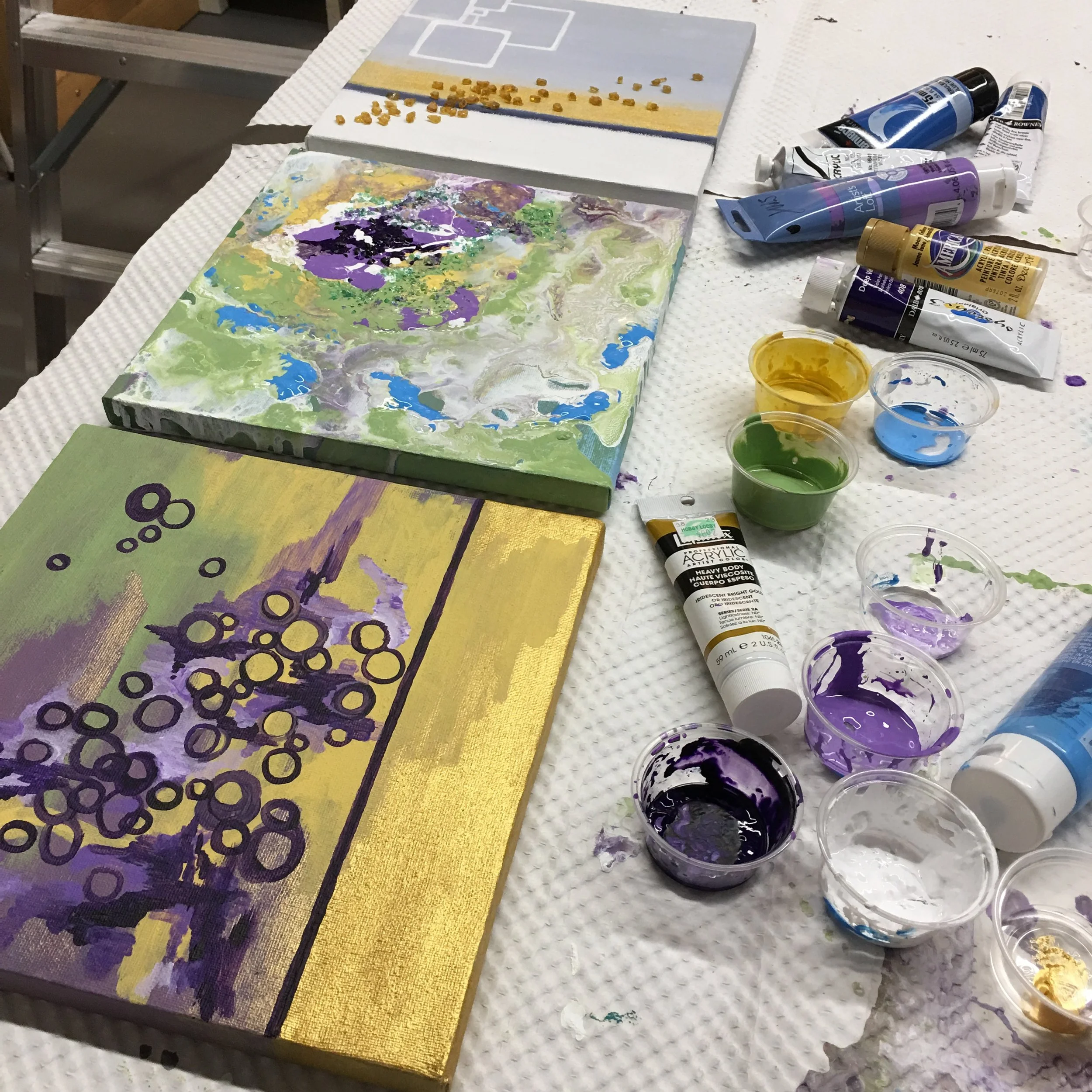 Two abstract paintings on a table with tubes of paint and small containers of mixed paint, some paint splatters on the table.