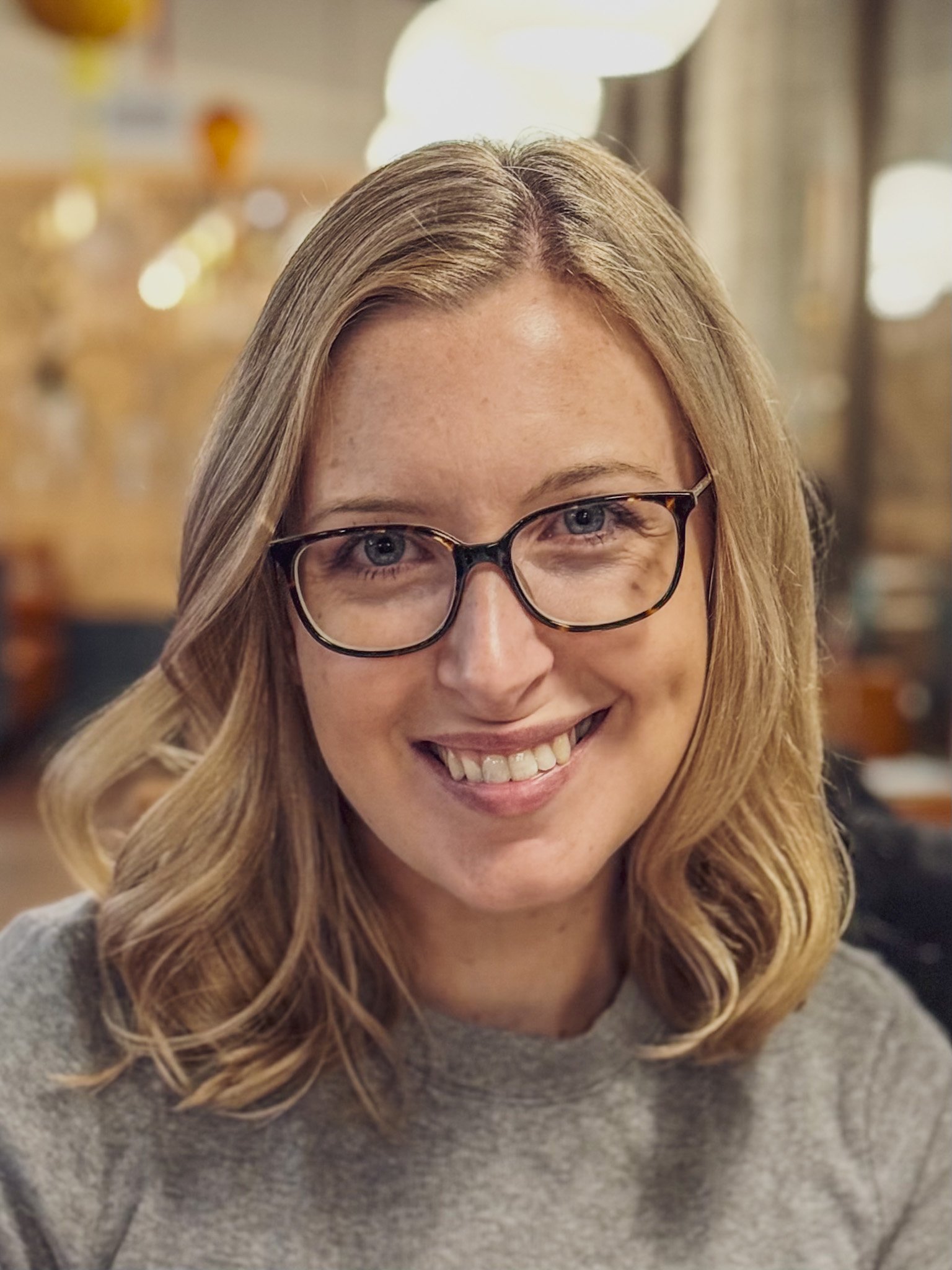 A woman with blonde hair, glasses, and a gray sweater smiling indoors.