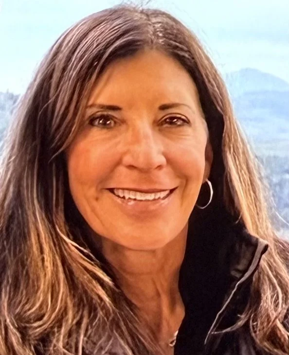 A woman with long brown hair smiling outdoors with mountains in the background.