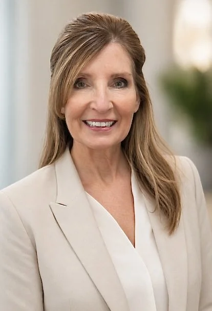 A smiling middle-aged woman with light brown hair styled with side-swept bangs, wearing a beige blazer over a white blouse, standing indoors with a blurred background.