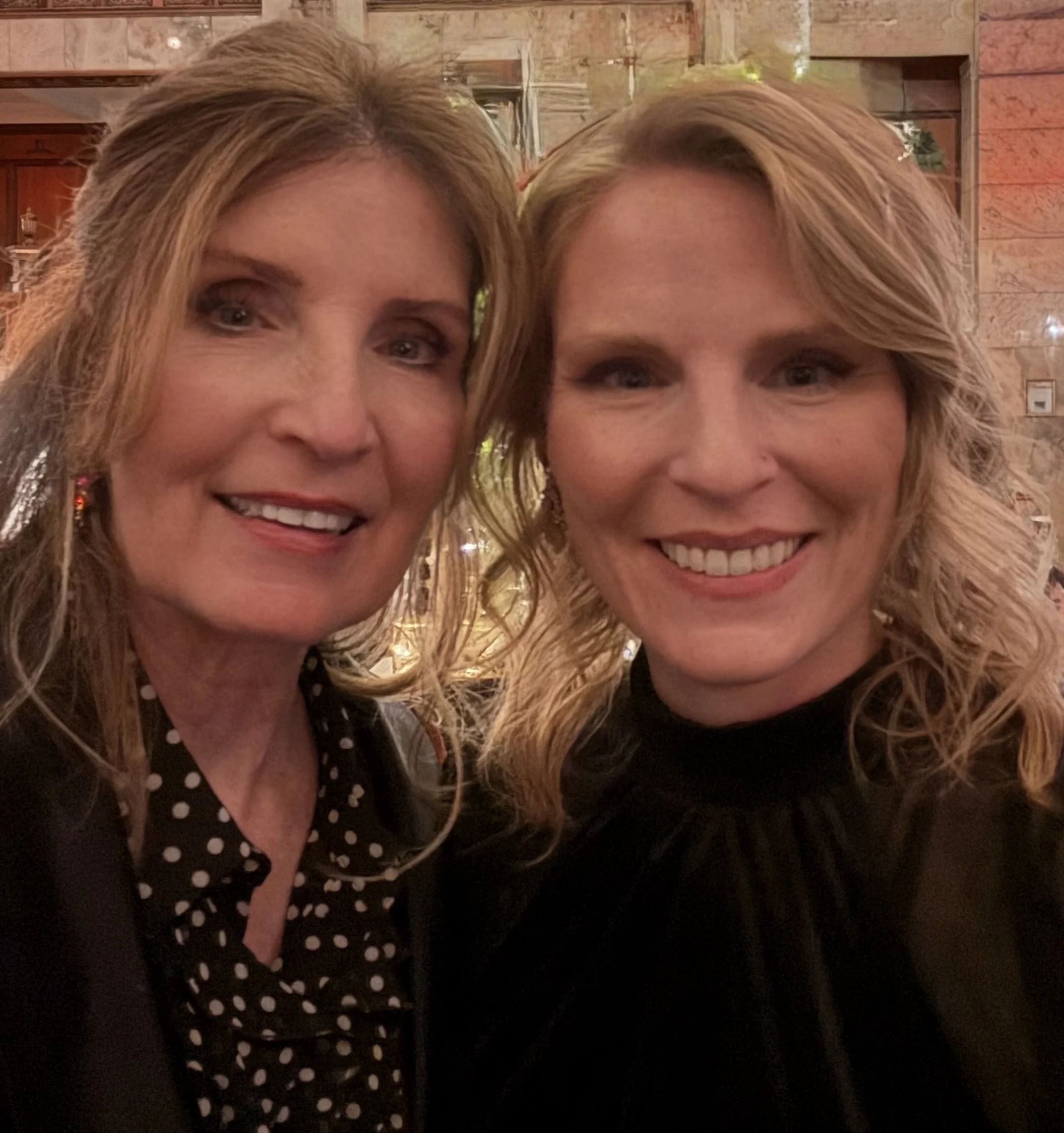 Close-up of two smiling women with blonde hair, one wearing a black top with white polka dots, in a warmly lit indoor setting with wooden and brick decor.