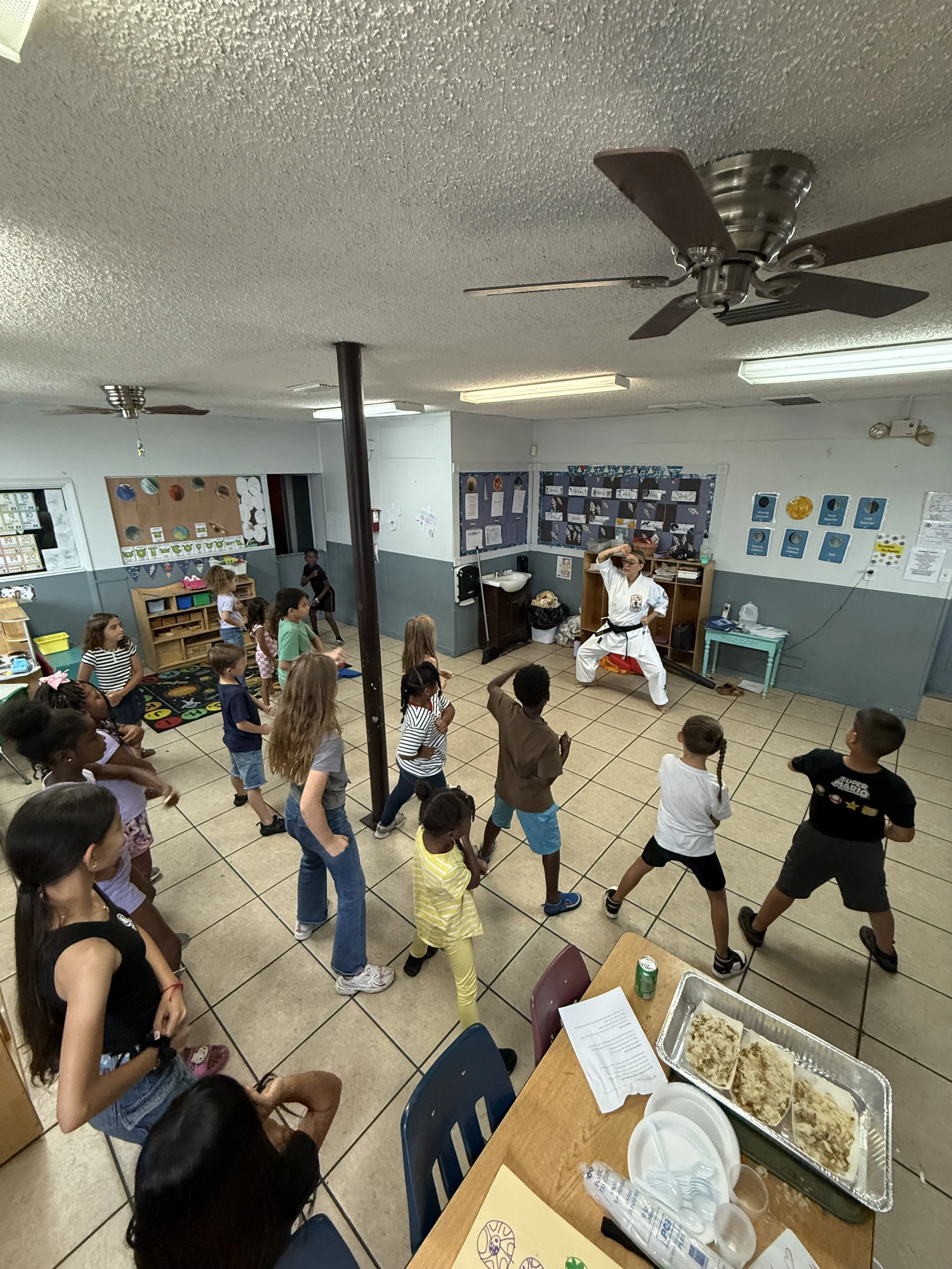 Children standing in a classroom, participating in an activity led by an instructor in a martial arts uniform. Tables with food, paper, and utensils are in the foreground.