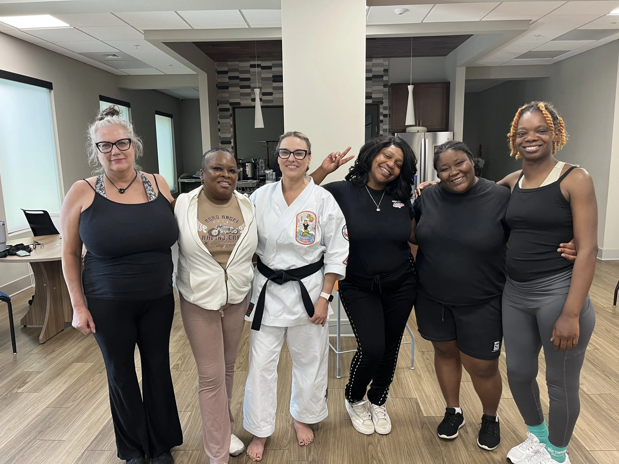 Group of six women standing together in a room, smiling and posing for a photo. Some women are wearing casual clothing, while one is dressed in a martial arts uniform. The room has modern decor with pendant lights and large windows.