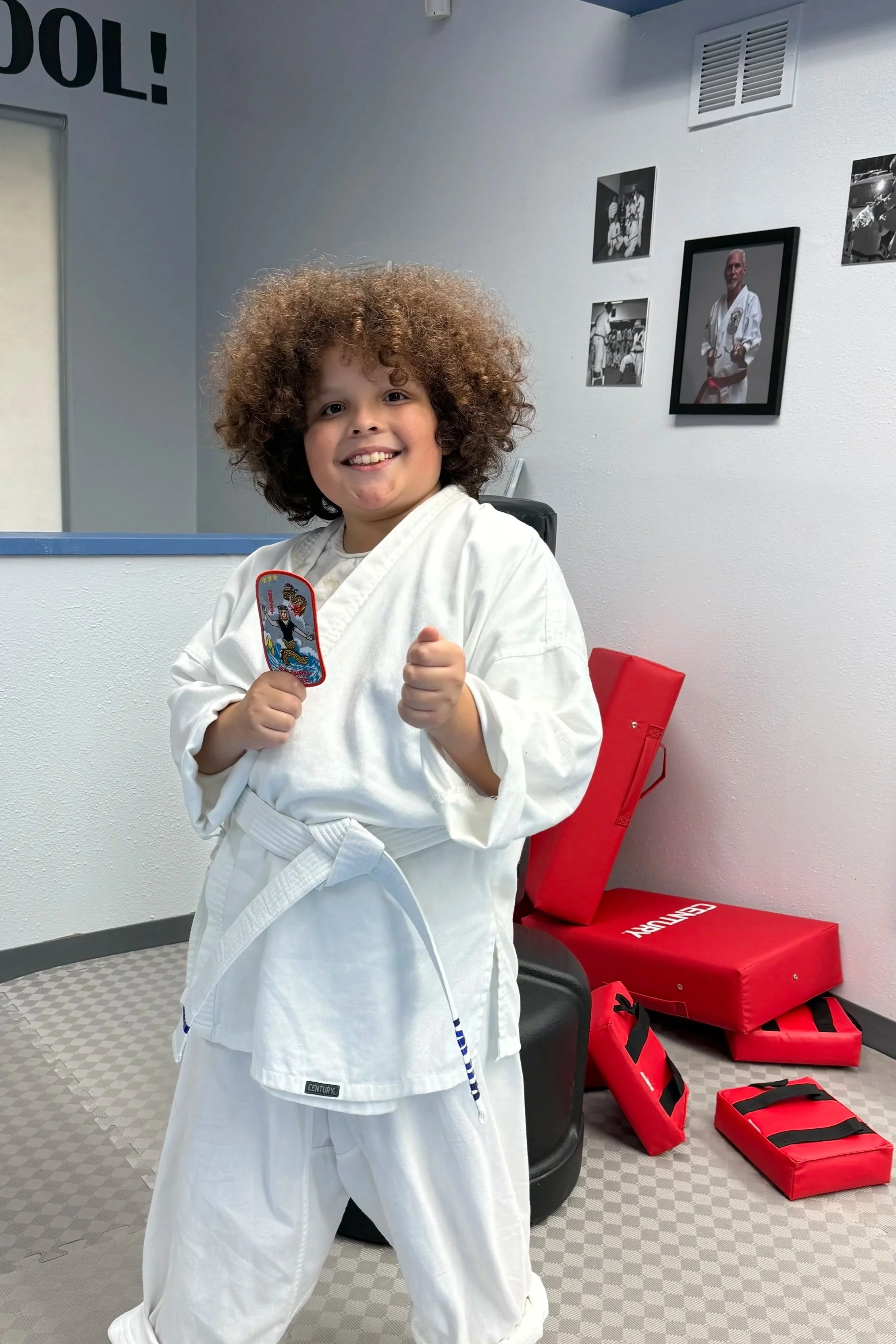A young child with curly hair wearing a white karate gi and a brown belt, smiling and giving a thumbs-up in a martial arts training room with red padded equipment behind them.