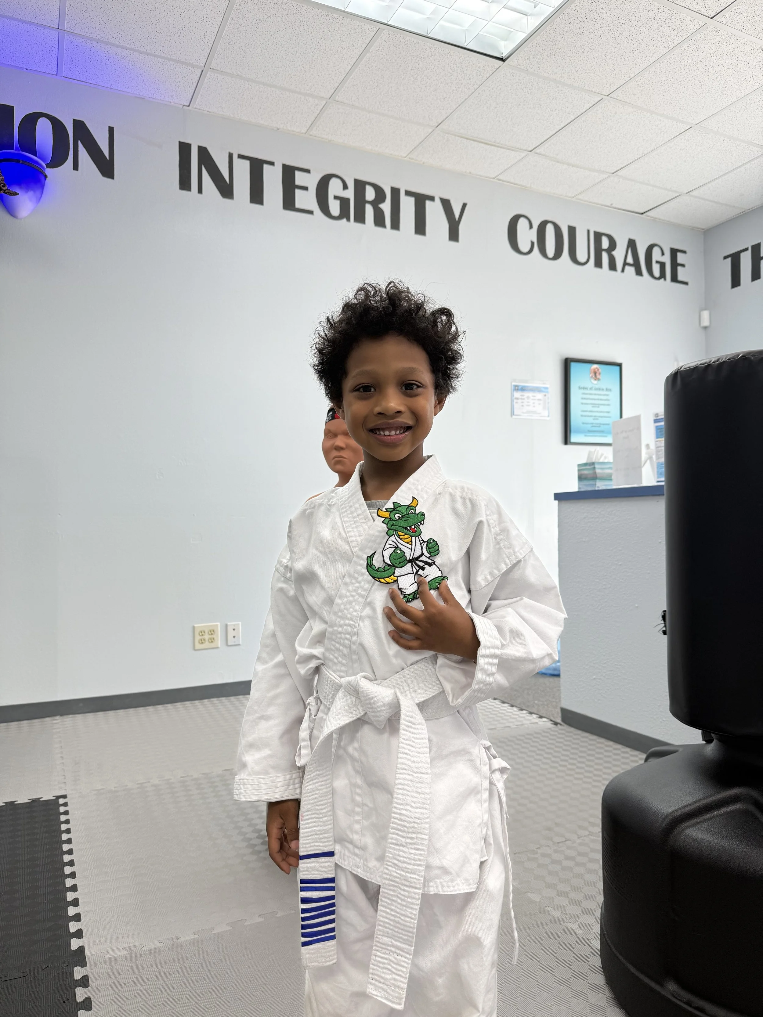 Young boy in a white martial arts uniform with a dragon patch on the chest, standing in a dojo. He is smiling and looking at the camera, with a person faintly visible in the background. The wall behind displays words: 'INTEGRITY' and 'COURAGE'.