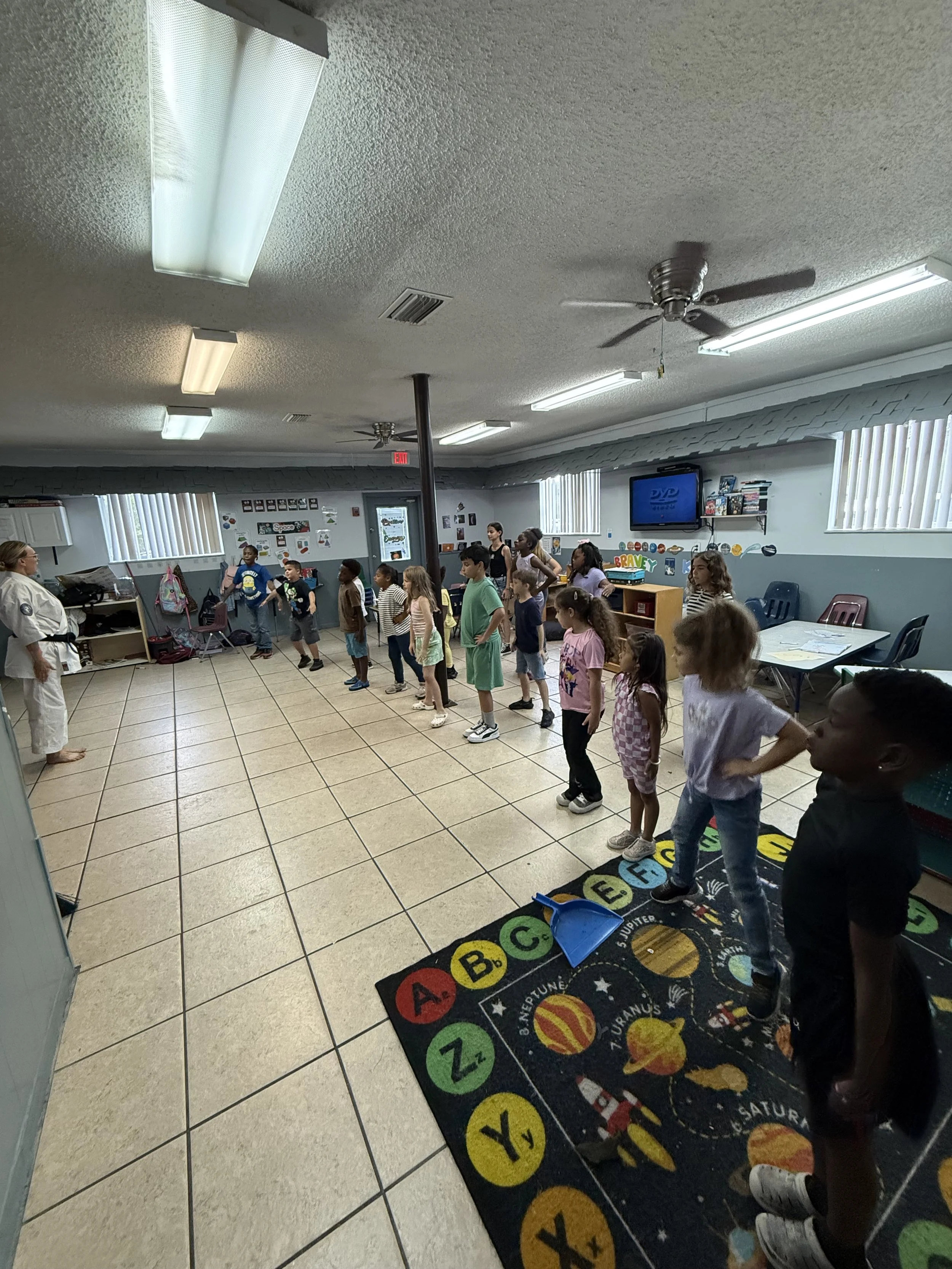 A group of children standing in a line in a classroom or activity room with some adults, likely participating in a group activity or game. The room has white walls, windows with blinds, ceiling lights, and posters on the wall. There is a colorful alphabet-themed rug on the tile floor.
