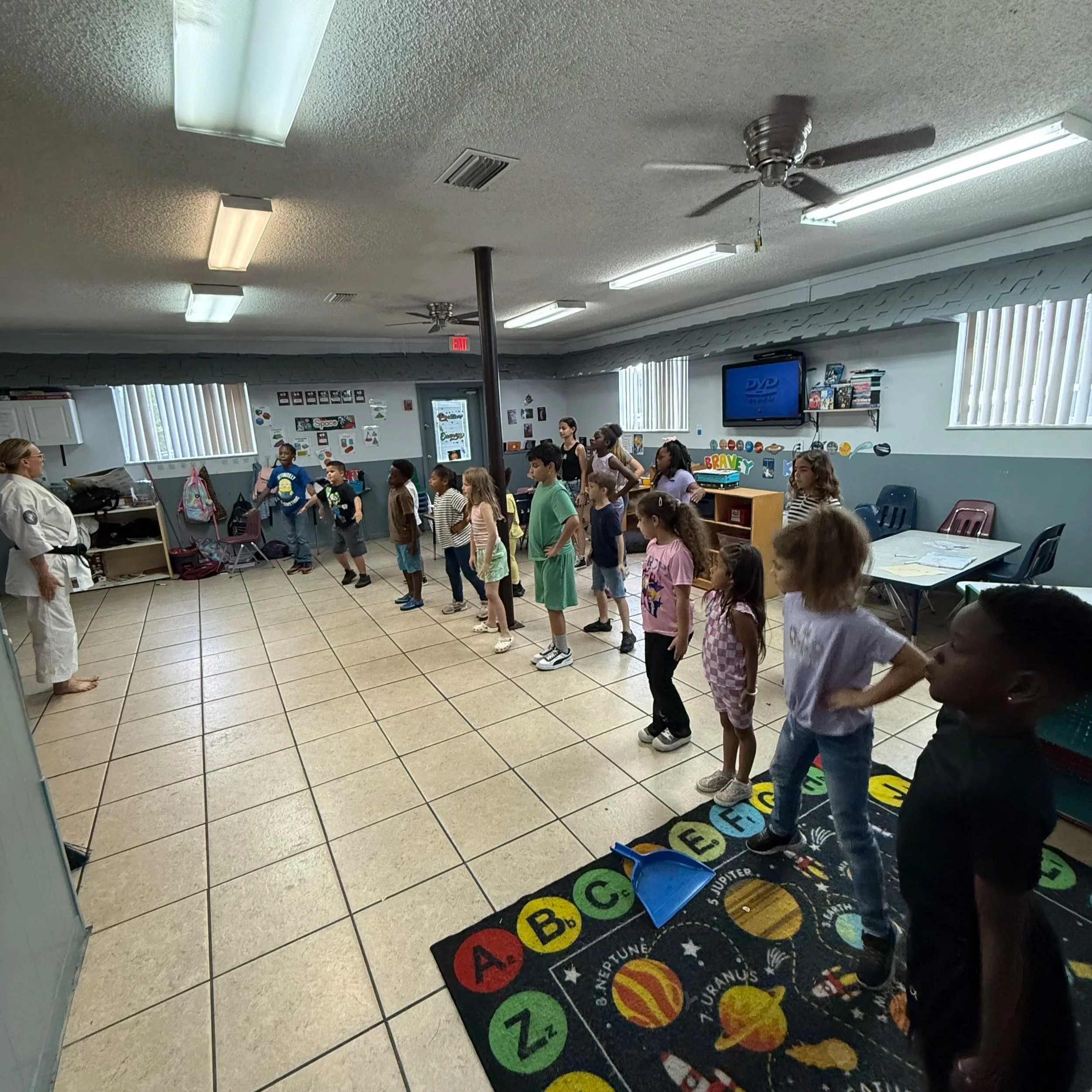 A classroom with children standing in line, a teacher at the front, a TV screen on the wall displaying the DVD logo, and educational posters and decorations on the walls.