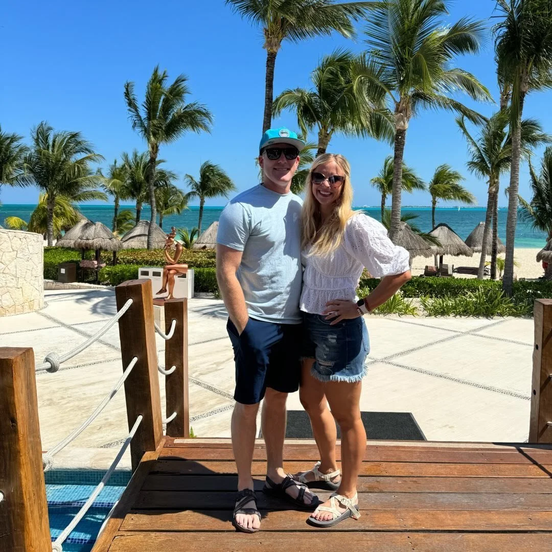 A couple posing on a wooden deck at a tropical beach resort with palm trees, thatched umbrellas, and the ocean in the background under a clear blue sky.