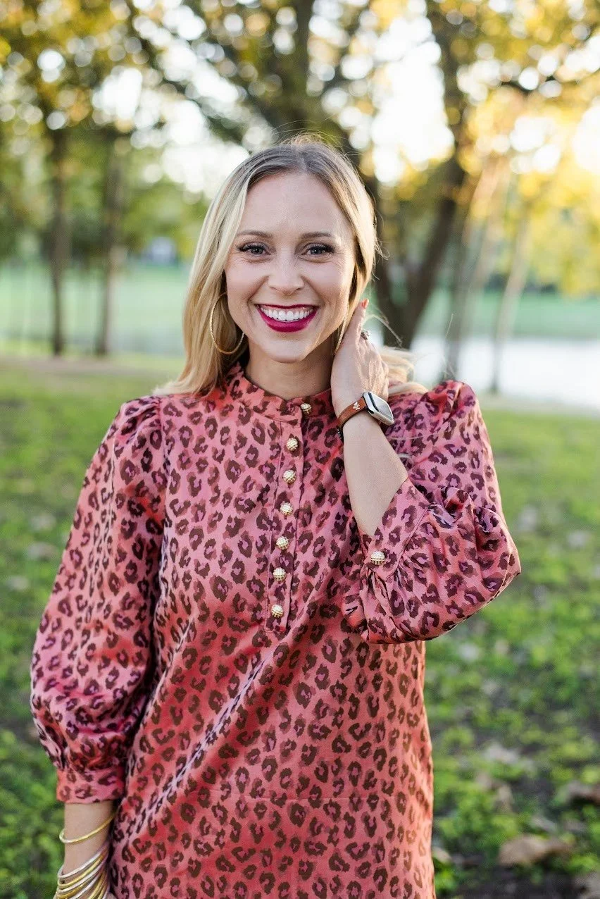 A smiling woman with blonde hair, wearing a pink leopard print blouse, gold jewelry, and a smartwatch, standing outdoors in a park with green grass and trees in the background.