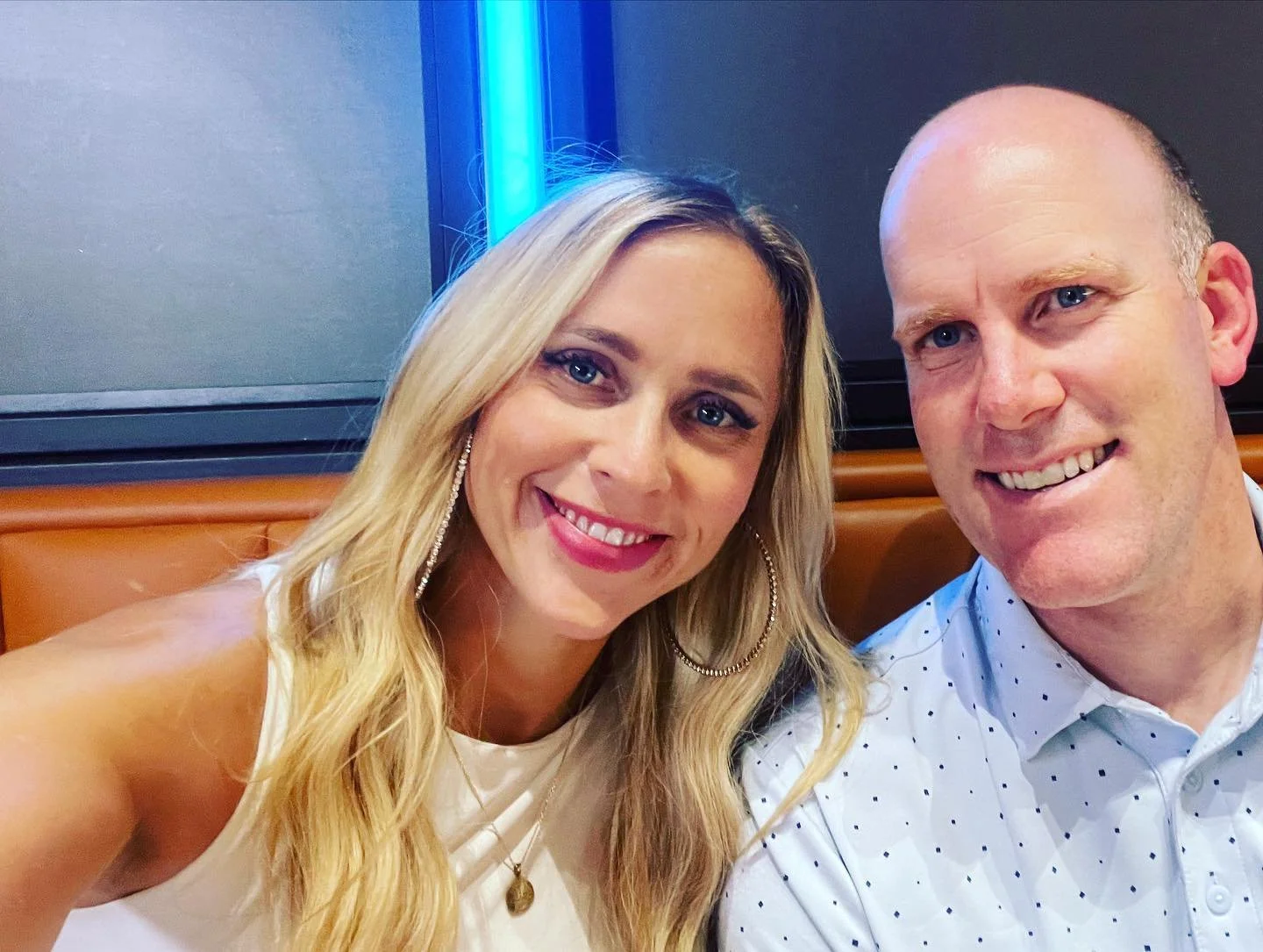 A smiling woman with blonde hair and a man with a shaved head taking a selfie together in an indoor setting, with blue lighting in the background.