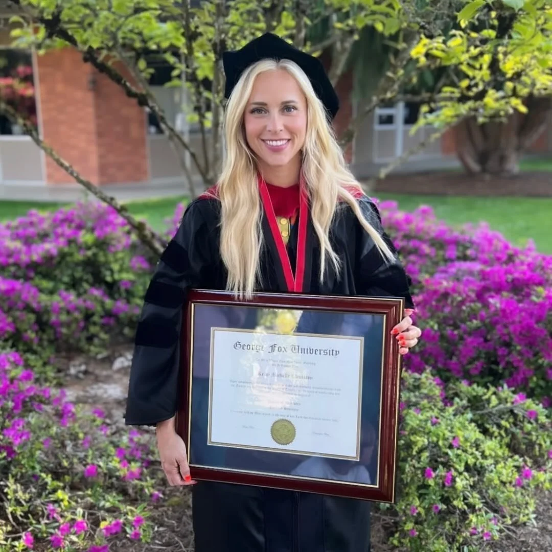 A young woman in academic regalia holding a framed diploma outside in front of pink flowers and green trees, smiling at the camera.