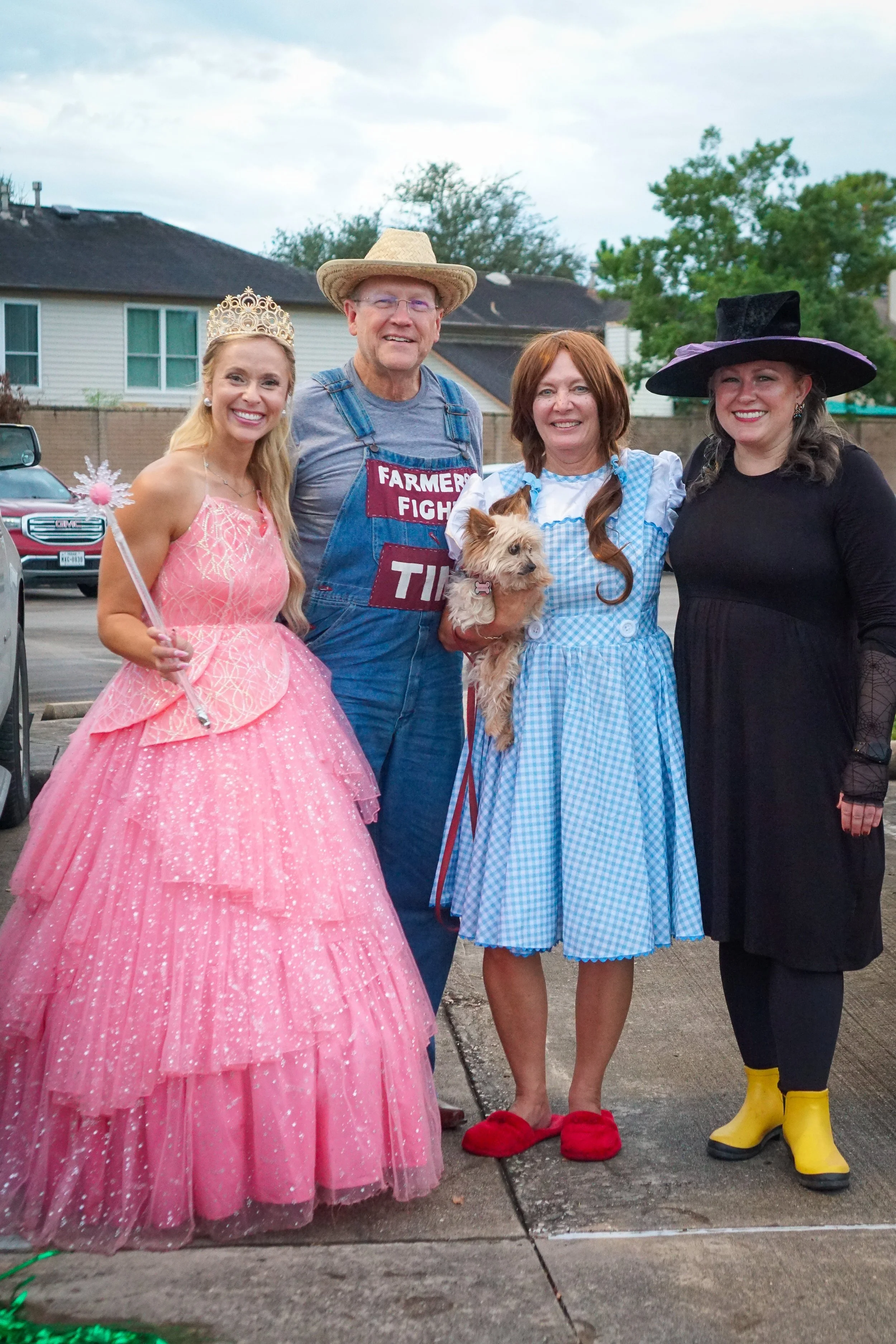 Four people in costumes standing outside with houses and trees in the background. The first person is a woman in a pink princess dress and tiara holding a wand. The second person is a man in overalls, a straw hat, and a shirt with a sign that says "Farmer Fight". The third person is a woman in a blue and white gingham dress holding a small dog. The fourth person is a woman in black with a large witch hat and yellow rain boots.