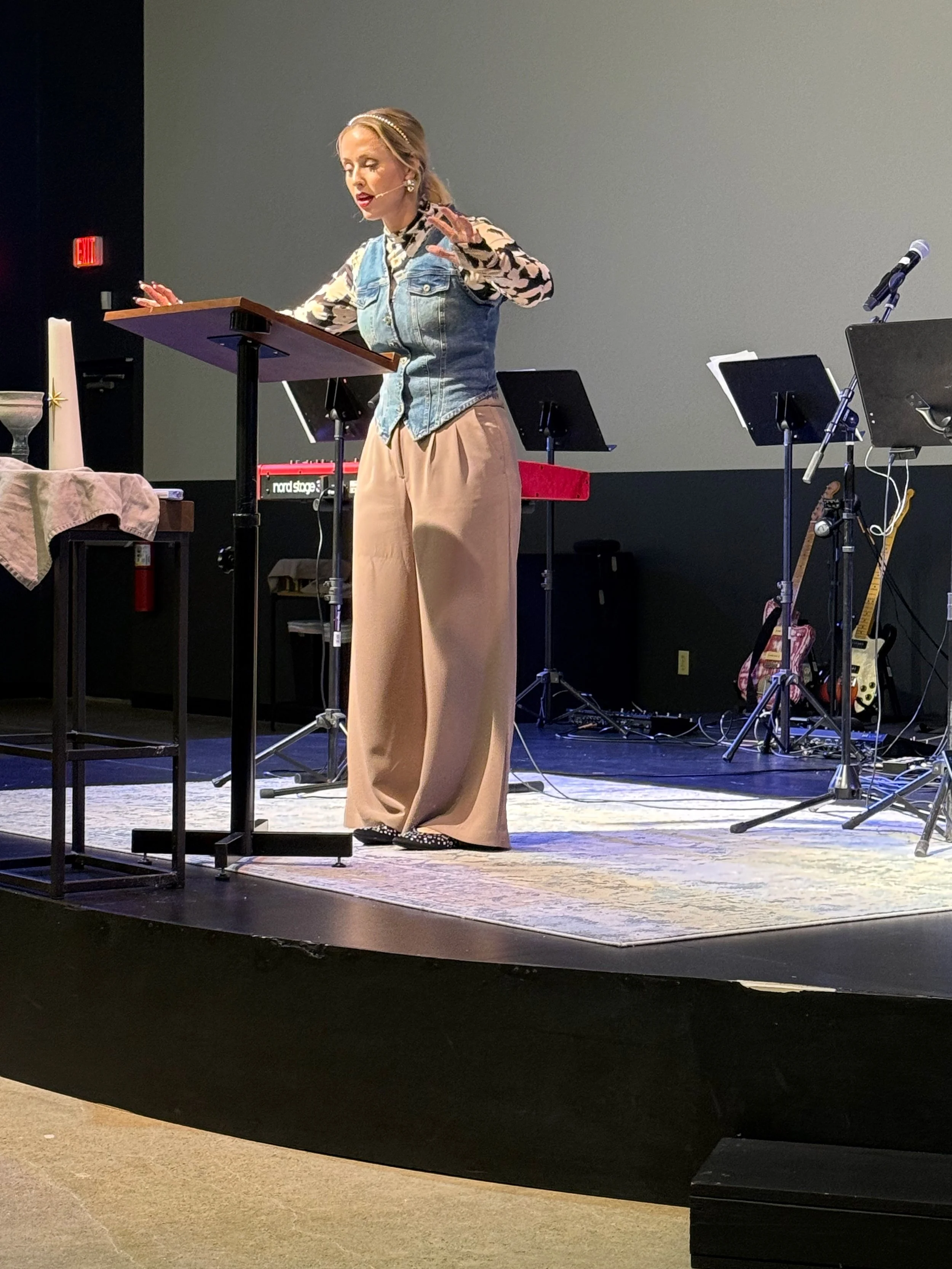 A woman dressed in a patterned blouse, denim vest, and wide-legged beige pants, standing on a stage at a podium, speaking with one hand raised. Musical instruments, music stands, and a microphone are visible on stage.