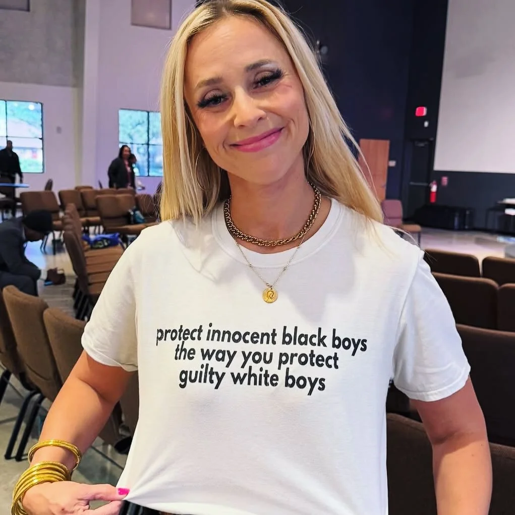 A woman with blond hair smiling and wearing a white T-shirt with a statement about protecting black boys and criticizing protections for white boys, in a large room with chairs and other people in the background.