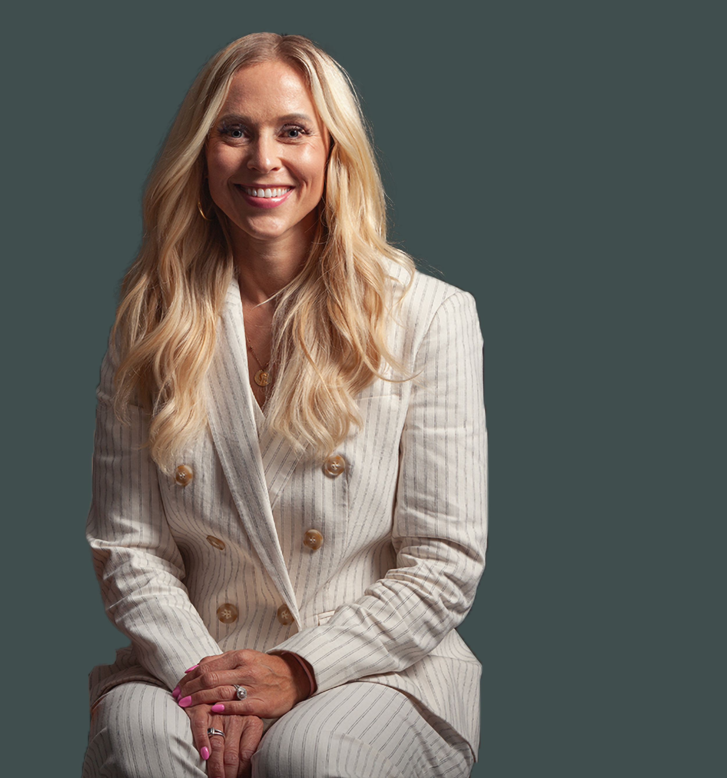 A woman with long blonde hair wearing a white pinstripe suit with large buttons, sitting with her hands clasped in her lap, smiling at the camera against a dark gray background.