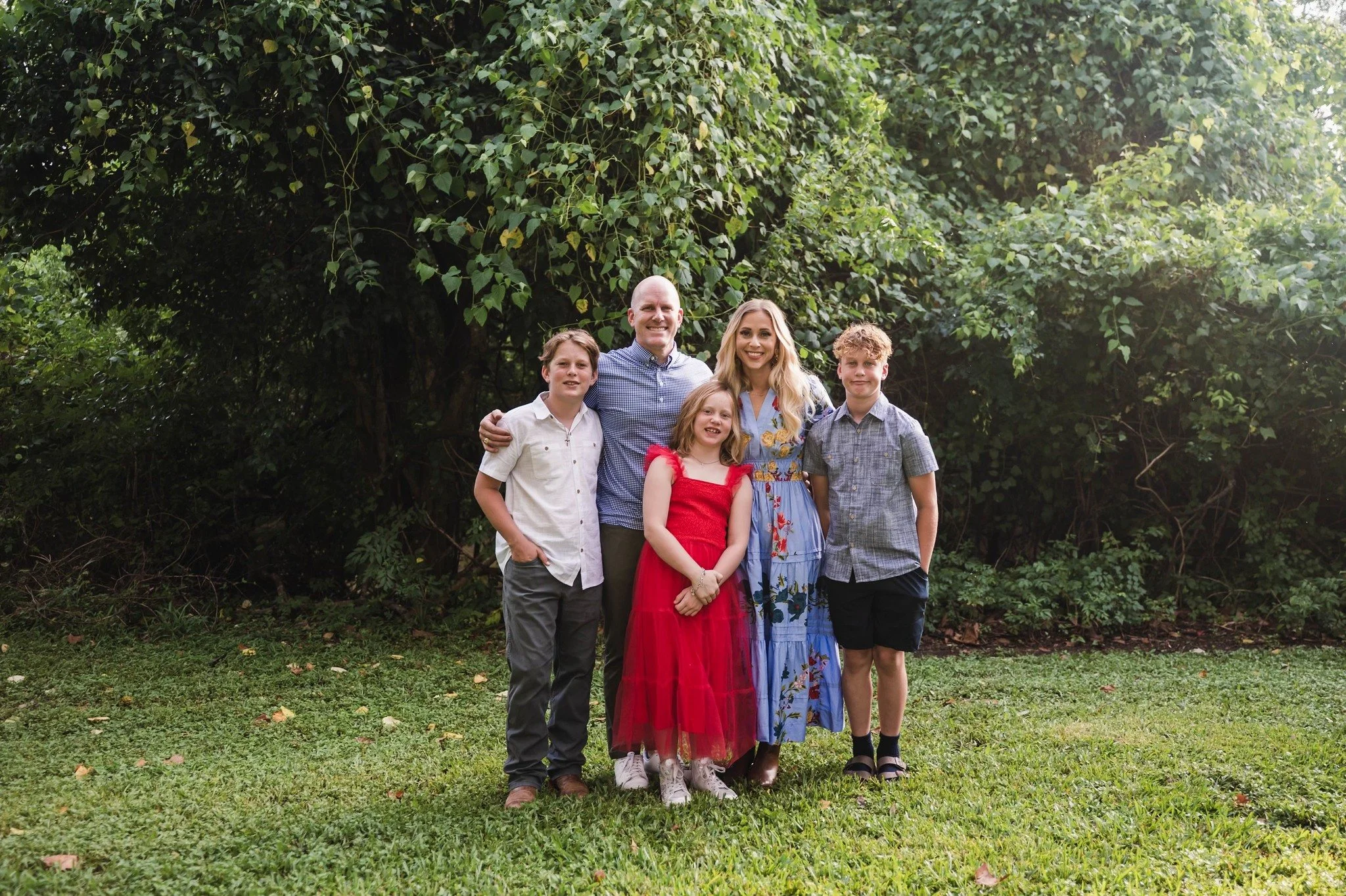 Family of five standing outdoors on grass near dense green foliage, smiling at the camera.