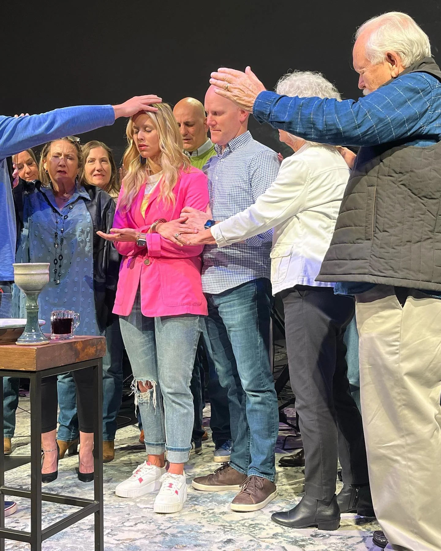 A group of people participating in a spiritual or prayer gathering. Some have eyes closed, hands raised or on their chests, with a person in the center receiving a blessing or prayer from others.