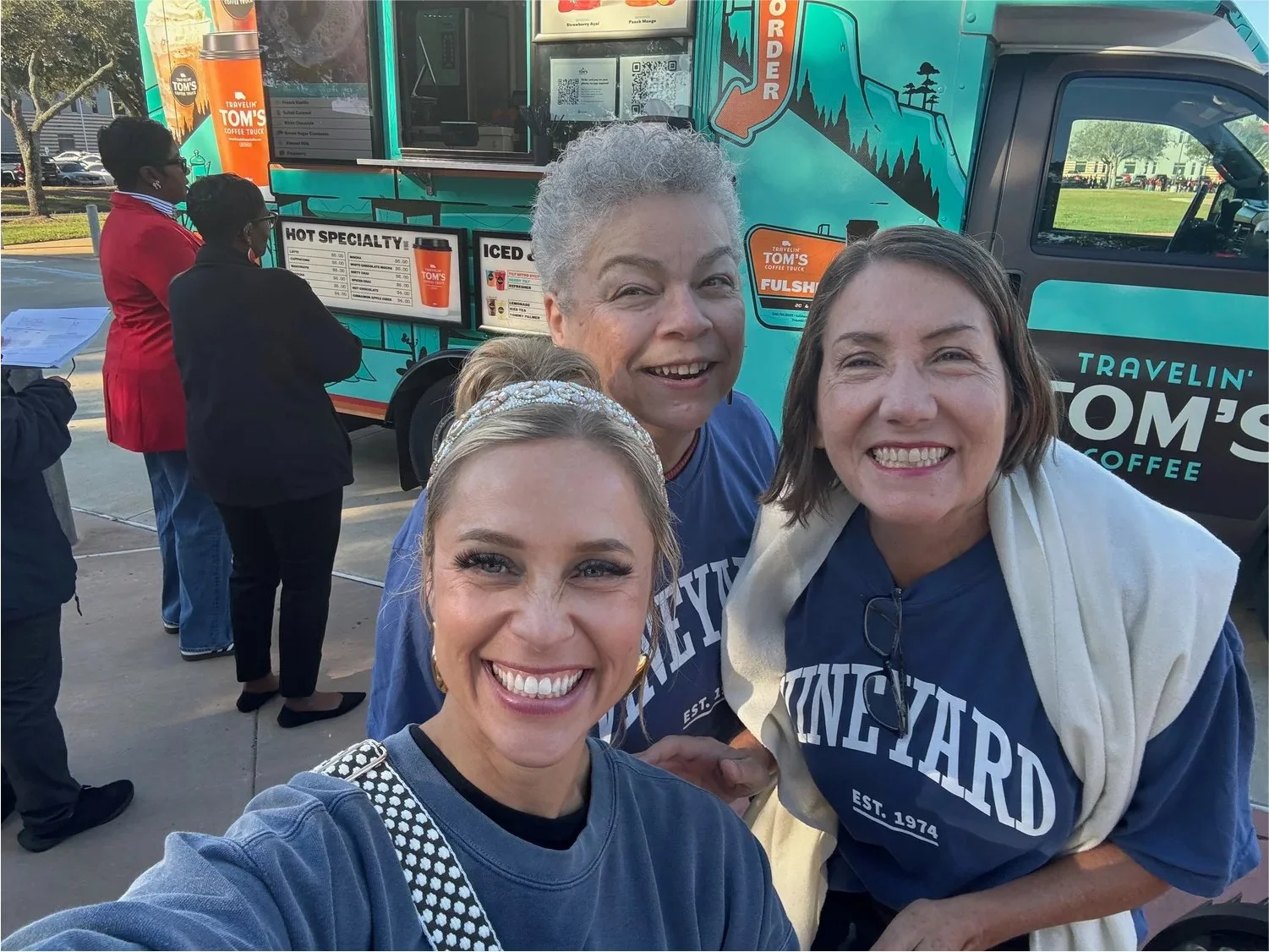 Three women smiling and taking a selfie in front of a food truck. There are two women in the background waiting in line. The food truck has a colorful mountain design and the words "Travelin' Tom's Coffee" on it.