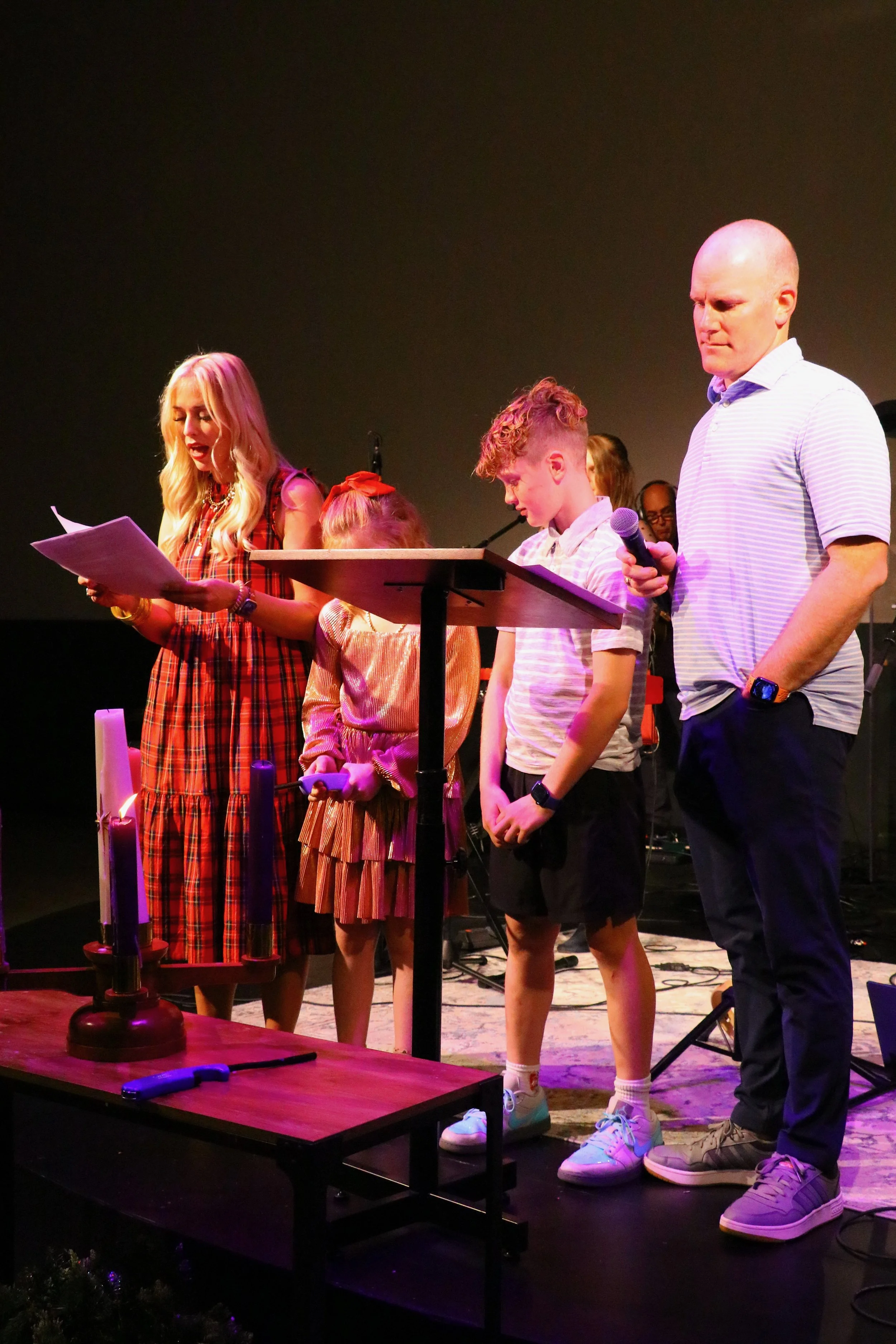 A group of people standing on stage, engaged in a reading or performance. Four individuals, including three young people and one adult man, are at a music stand with sheets of paper. Candles are lit nearby, and microphones and musical equipment are visible in the background.