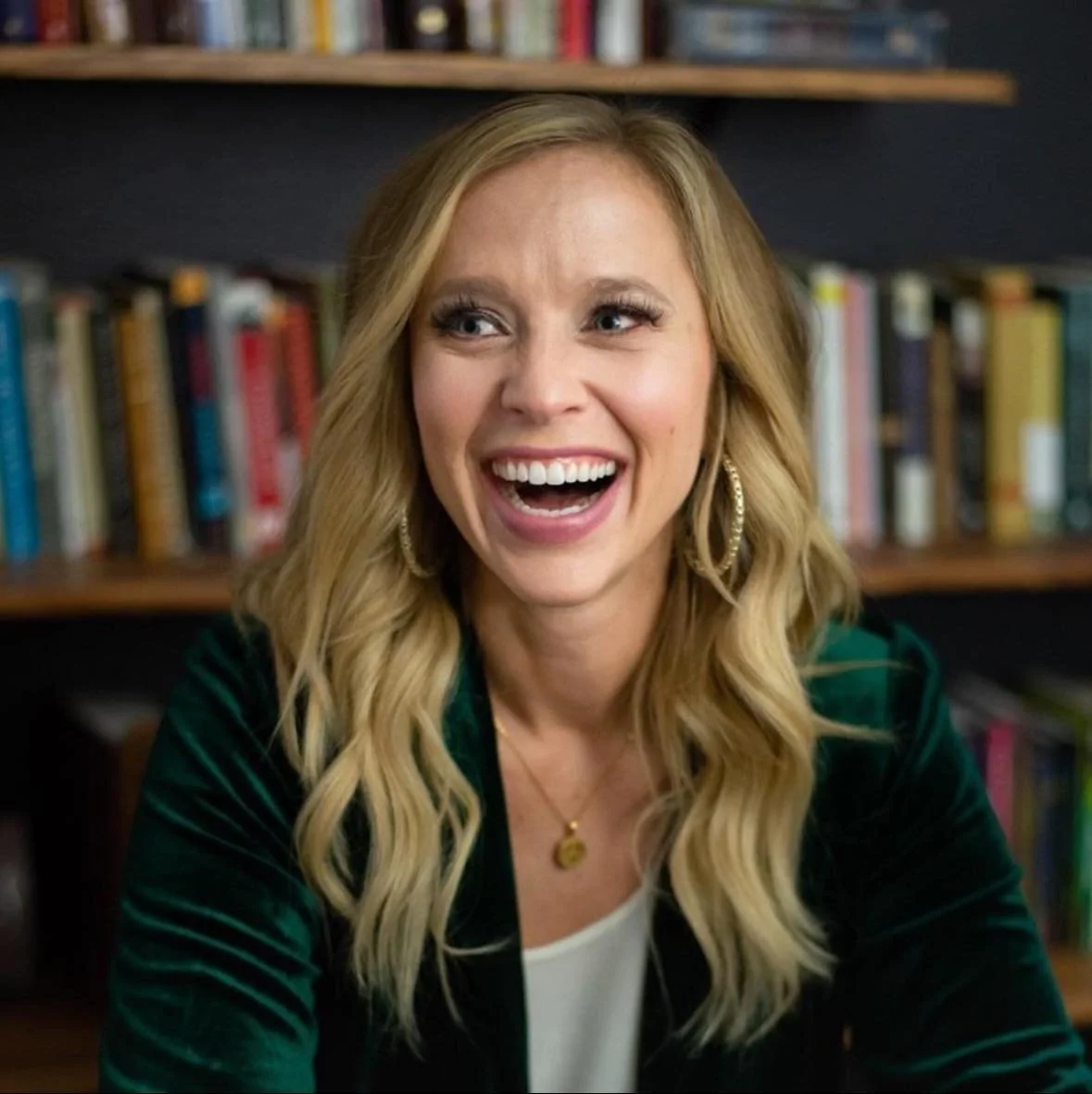 A woman with long blonde hair, wearing hoop earrings, smiling widely, indoors with a background of a bookshelf filled with books.