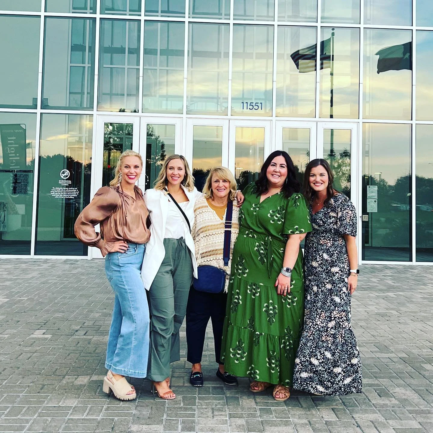Five women standing together outside in front of a large glass building, smiling at the camera, during sunset.