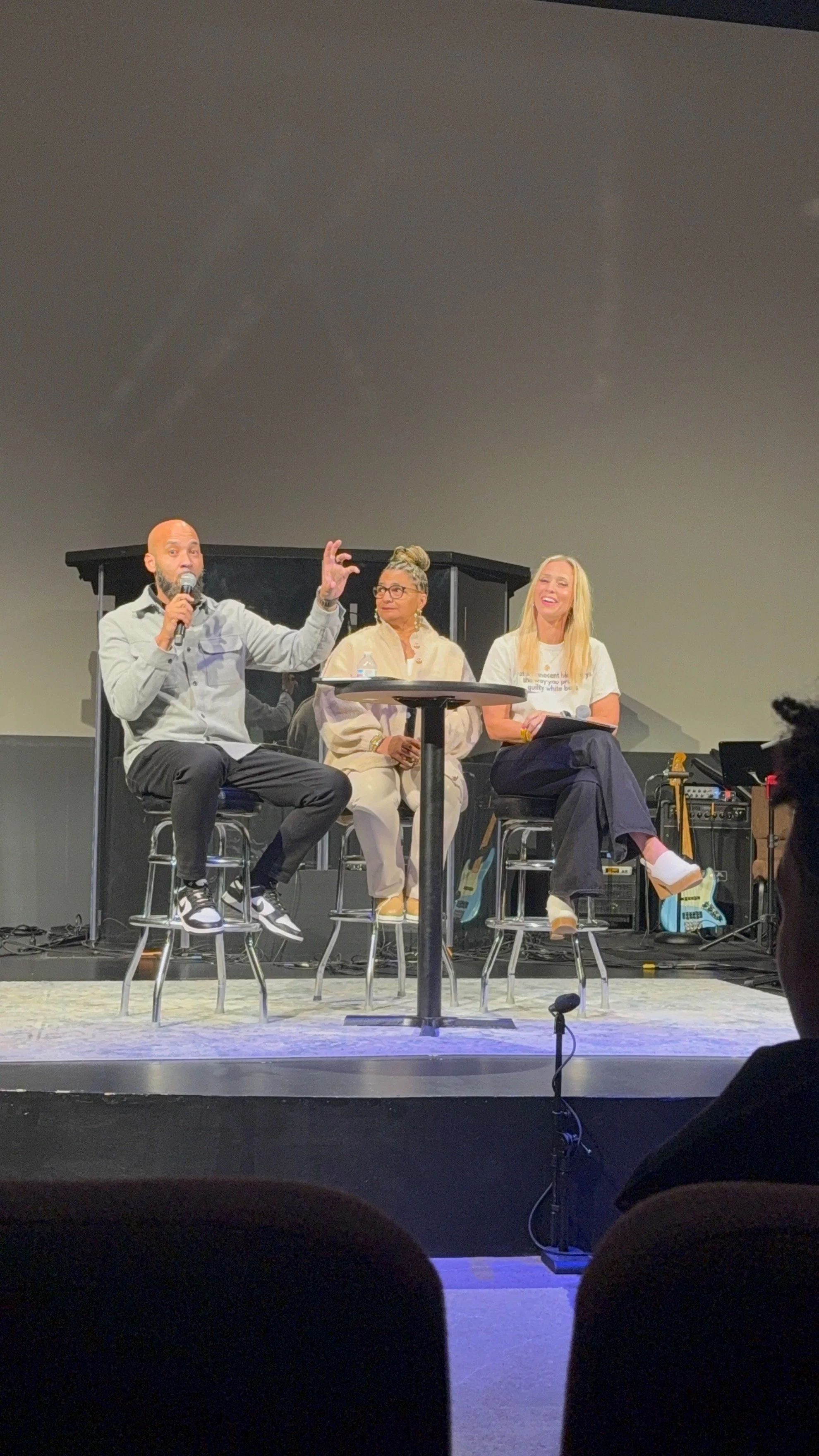 A panel of three people sitting on stools on a stage, engaging in a discussion or conversation. One person is speaking into a microphone, while the others listen. There are musical instruments and equipment behind them.