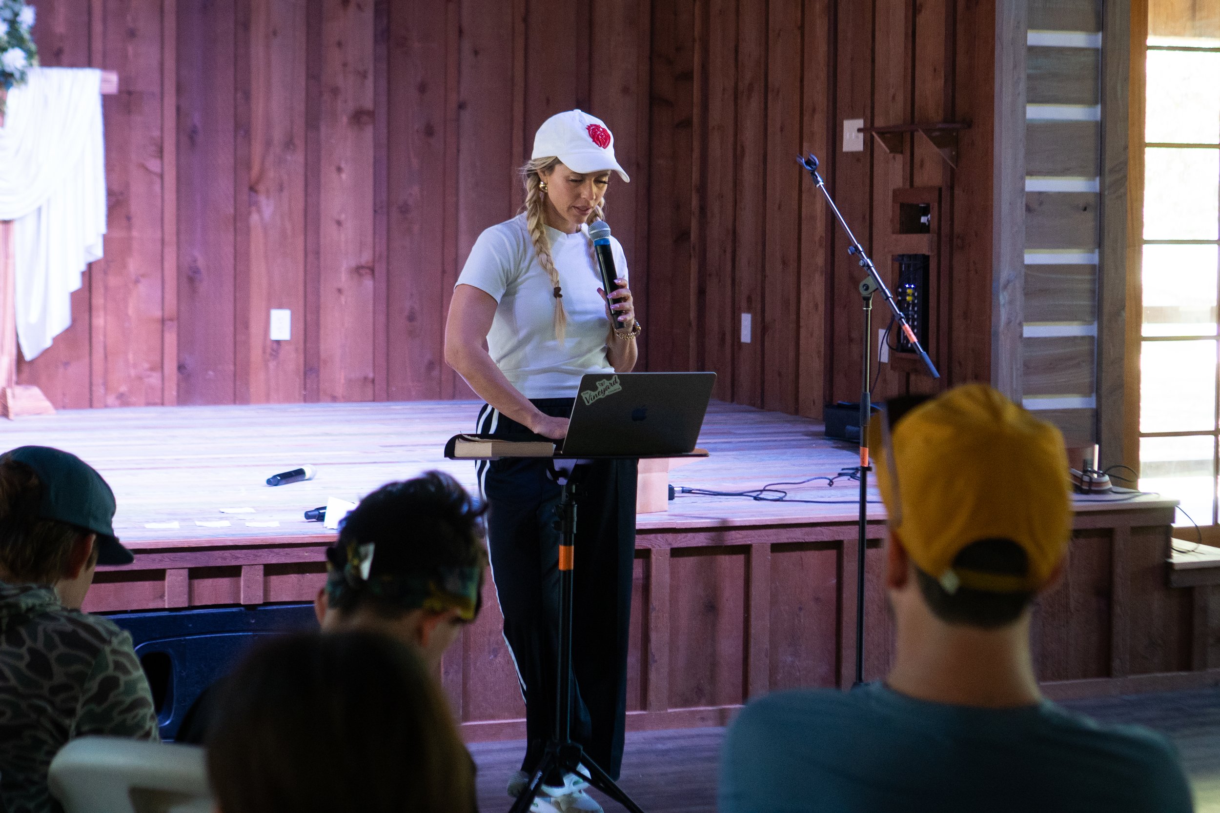 A woman with blonde braids, wearing a white cap with a red emblem, white t-shirt, and black pants, stands on a wooden stage holding a microphone and looks at a laptop, while several children sit in the foreground, facing her. The stage has a wooden wall with vertical planks and a window that lets in natural light.