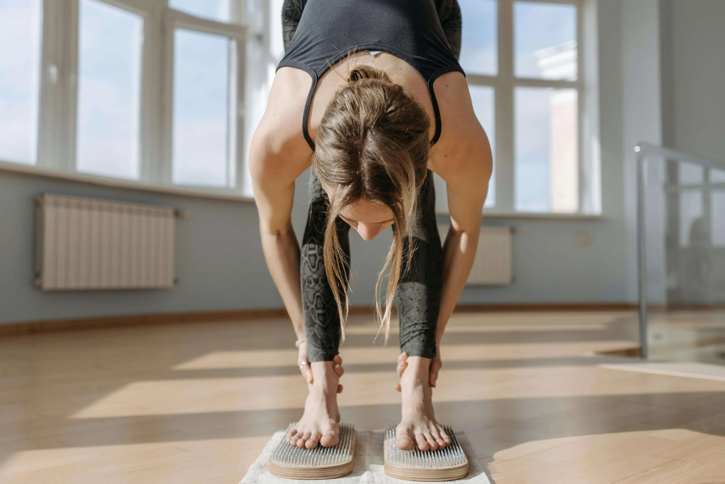 Woman practicing yoga in a bright room, standing in a forward bend position on a textured yoga mat.
