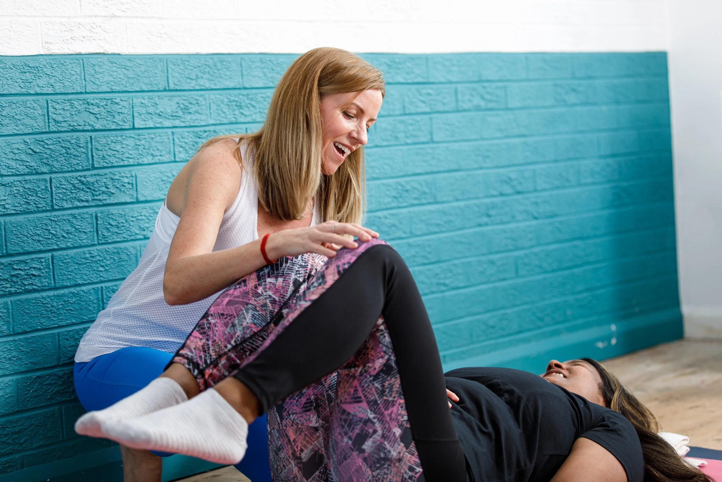 Two women engaging in a playful wrestling match on the floor, one standing and the other lying down, in front of a turquoise brick wall.