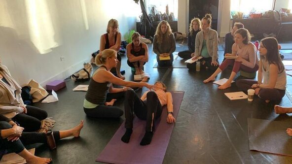 A woman demonstrating first aid techniques on a person lying on a yoga mat, surrounded by a group of people in a spacious room.