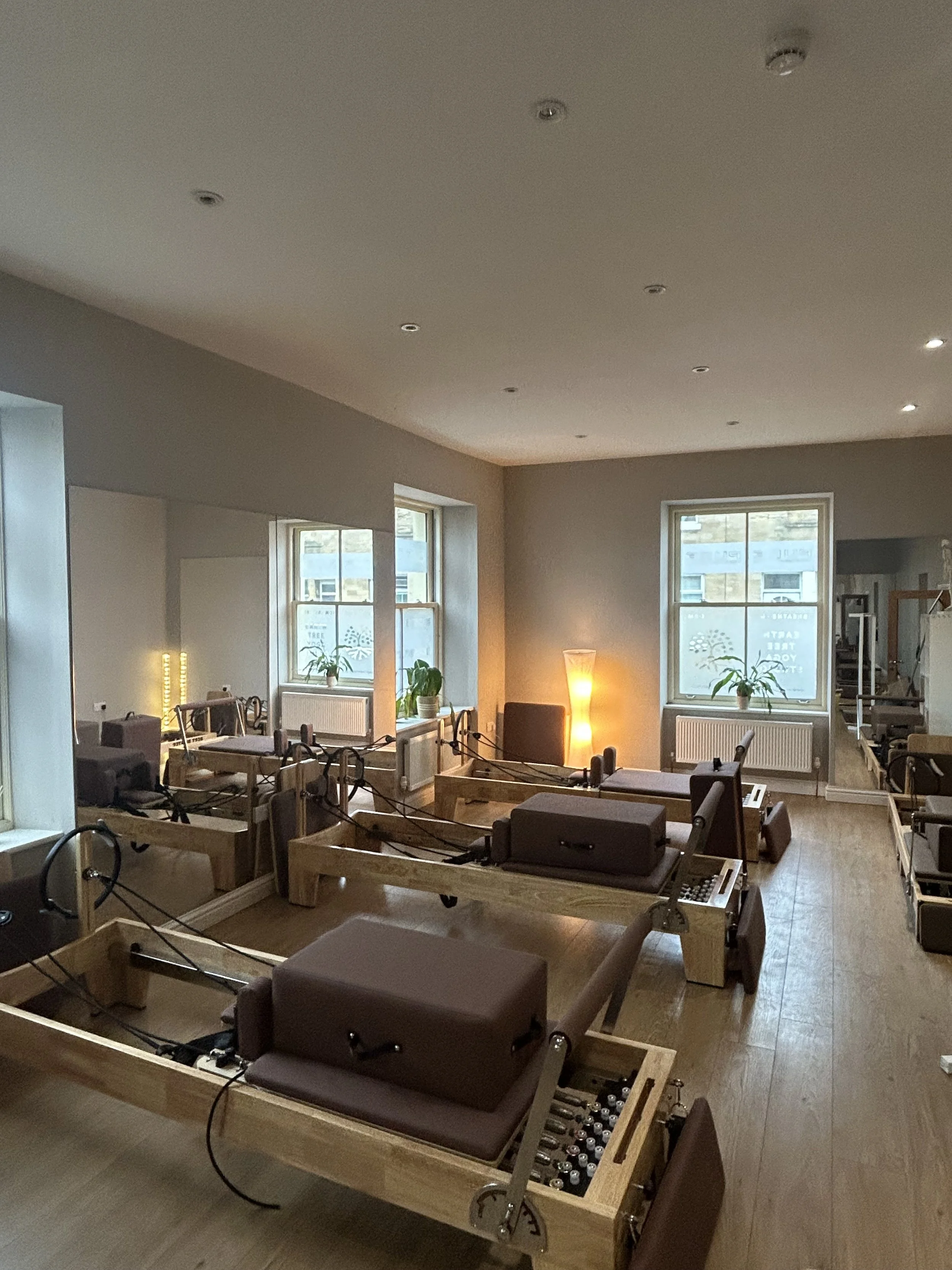 A room with wooden tables and chairs, some of which are connected to medical devices and monitoring equipment. The room has large windows letting in natural light, and some plants on the window sills. The ceiling has multiple small recessed lights and a smoke detector. The overall setup suggests a rehabilitation or therapy center.