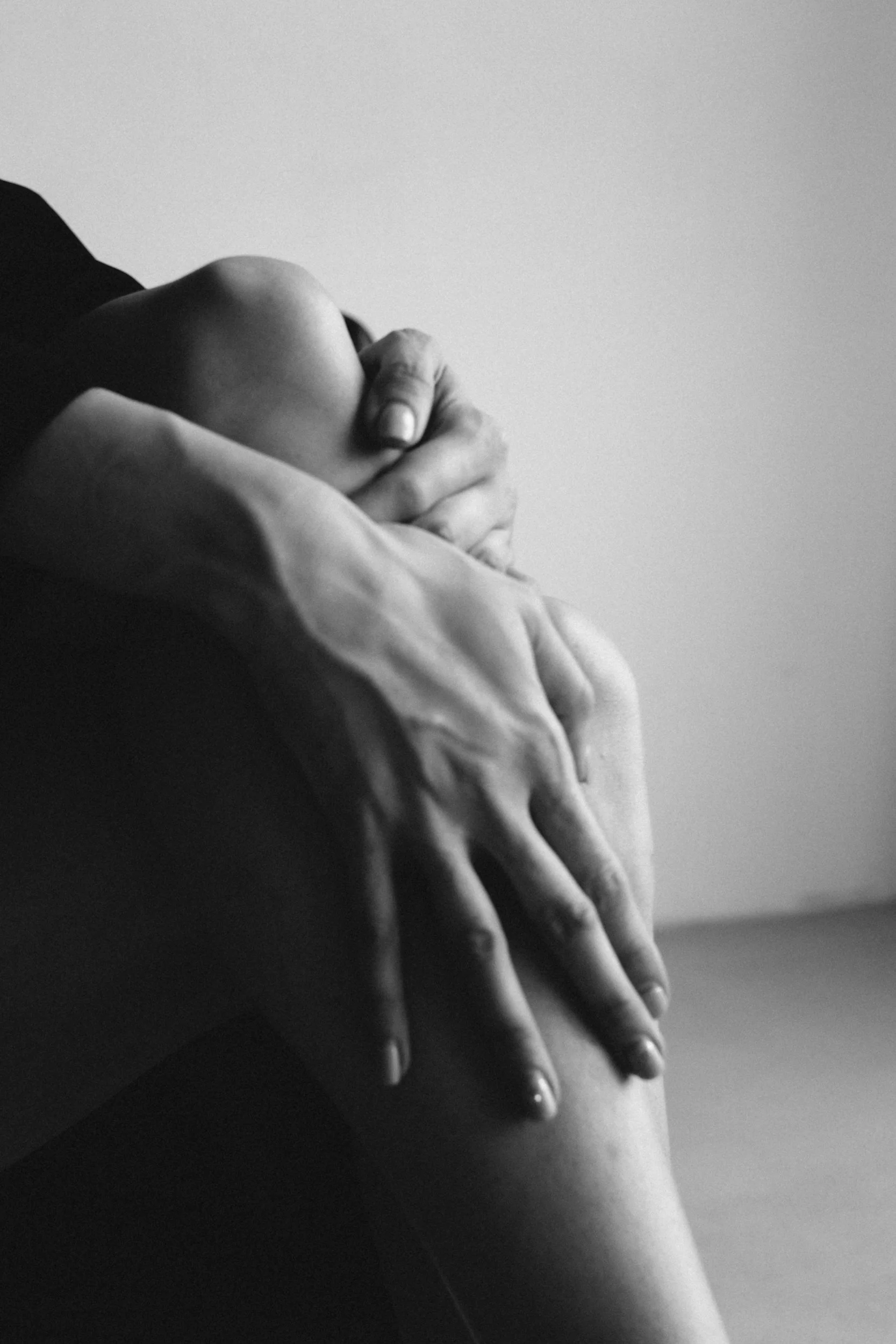 Close-up of a person's hand gently holding their knee, with a focus on the fingers and knee. The image is in black and white.