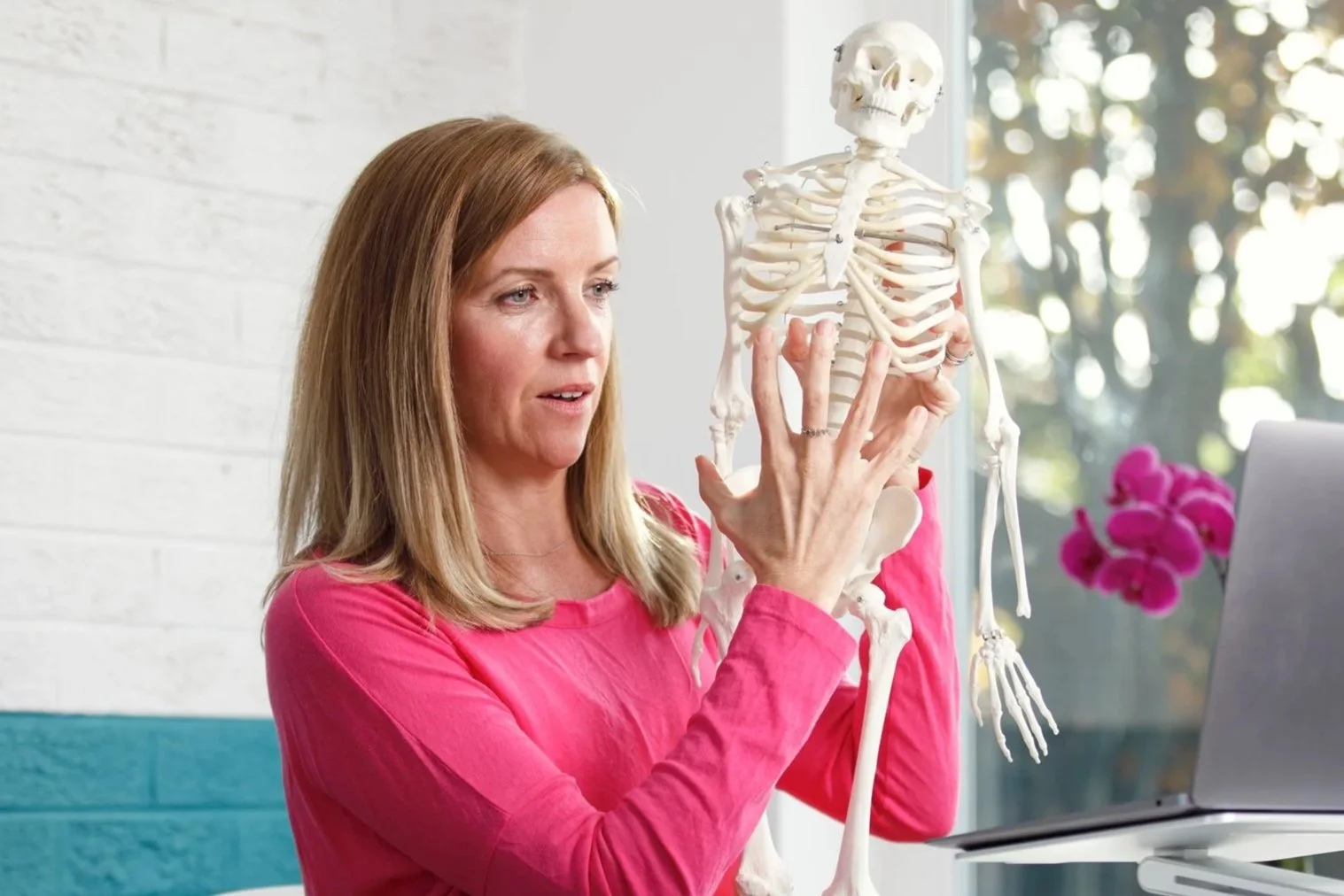 Woman in a pink shirt showing a human skeleton model to a computer.