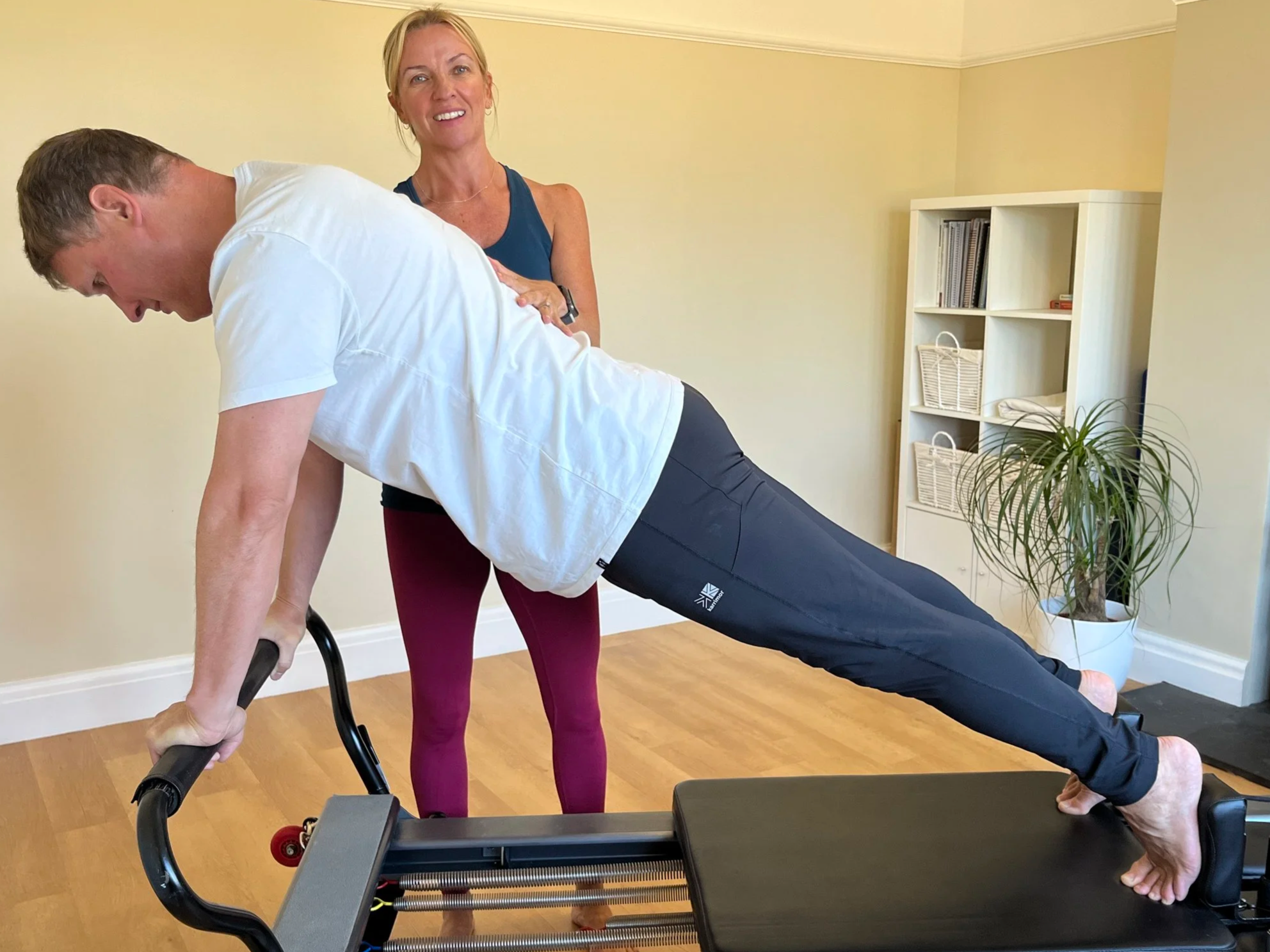 Louisa Thomas  guiding a man through a Pilates exercise on a reformer machine in a home studio, with a bookshelf and plant in the background.