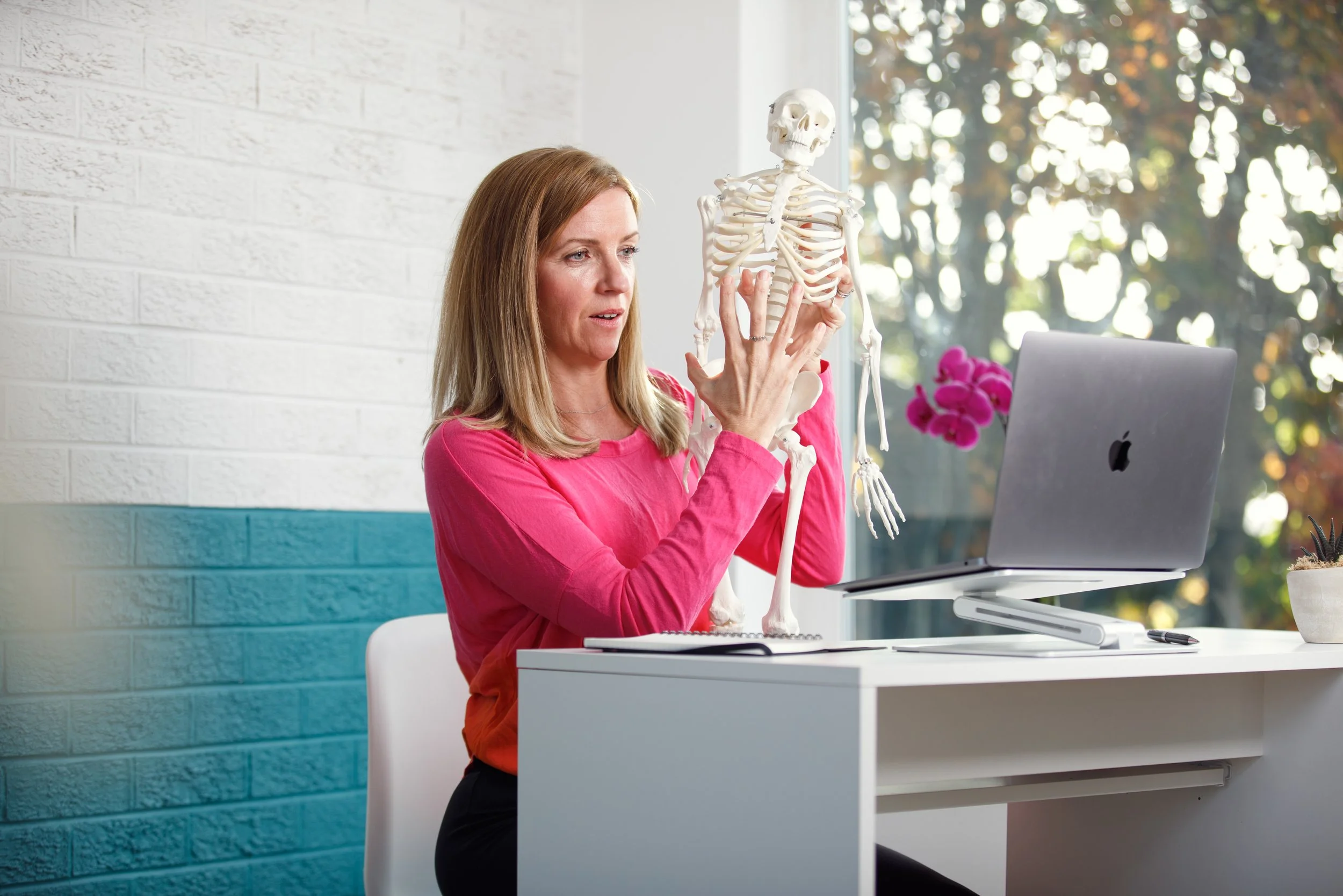 Louisa Thomas with blonde hair wearing a pink top sitting at a white desk, holding a skeleton model near her face while looking at her laptop with a plant and a window with trees in the background.