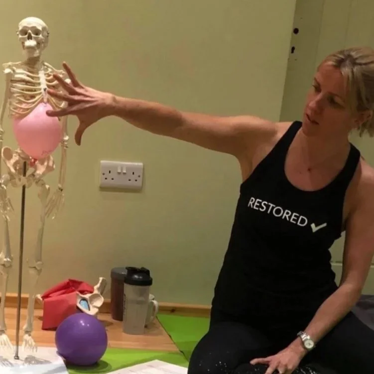 Louisa Thomas in a black tank top that says 'Restored' is stretching her arm to touch a skeleton with a pink balloon in its chest cavity. There are a purple exercise ball, a water bottle, and papers on a table nearby.