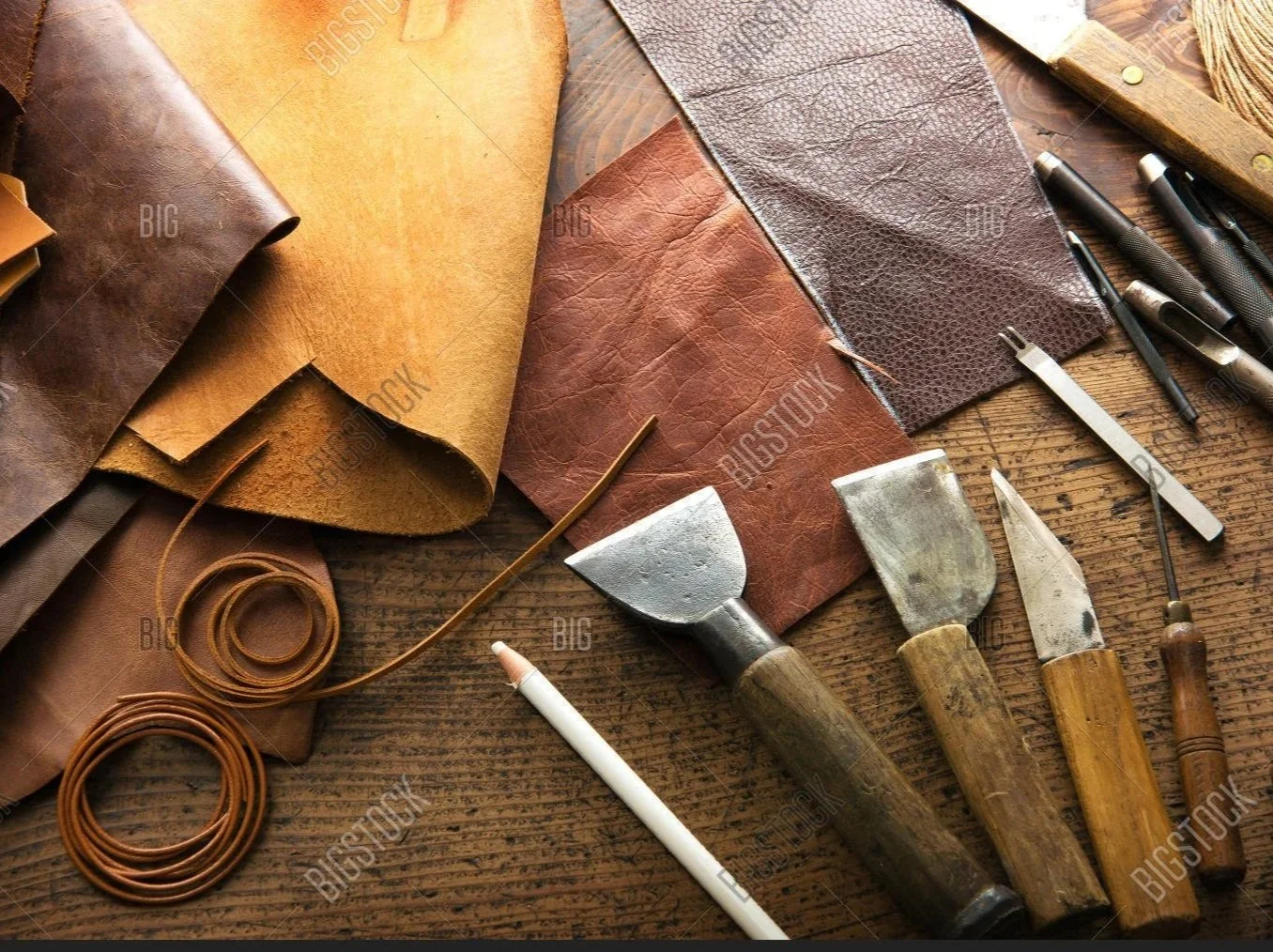 A variety of leatherworking tools and leather pieces on a wooden surface, including chisels, knives, pens, and rolls of leather in different shades of brown.