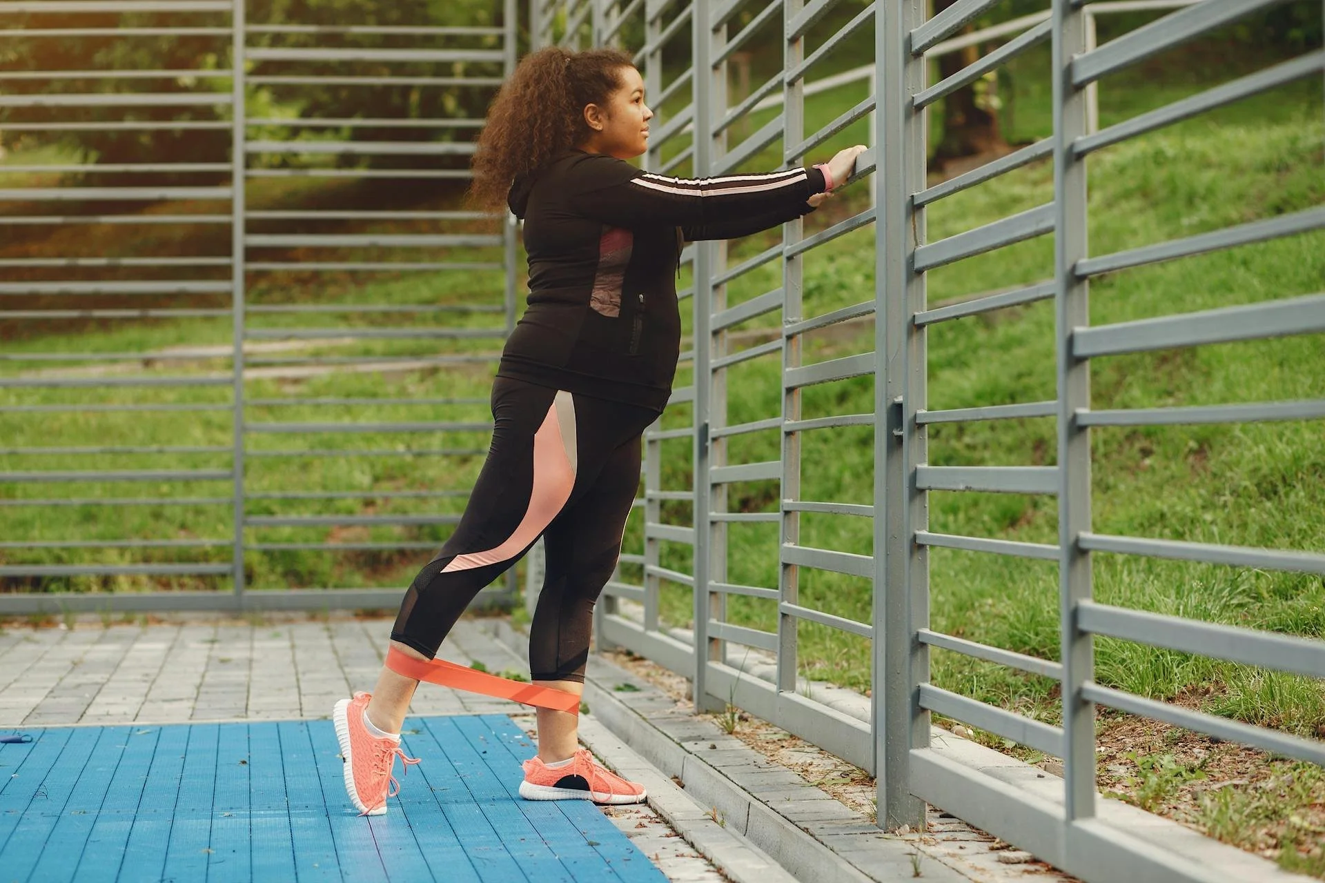 Woman exercising outdoors with resistance band around her ankles, stretching her legs against a metal fence.