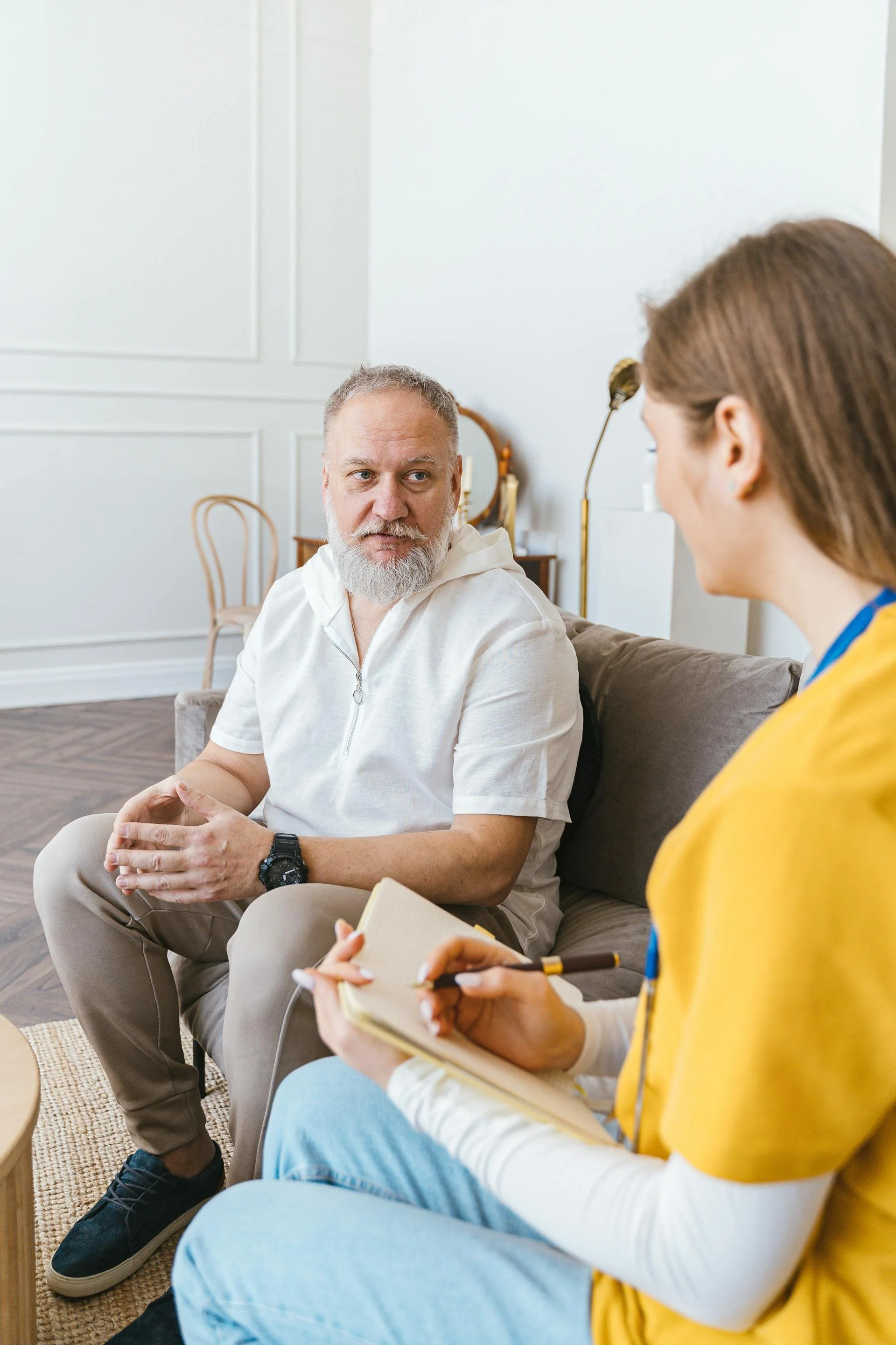A man with gray hair and a beard, wearing a white hoodie and beige pants, talking to a woman in medical scrubs holding a notepad and pen, in a room with white walls and wooden furniture.