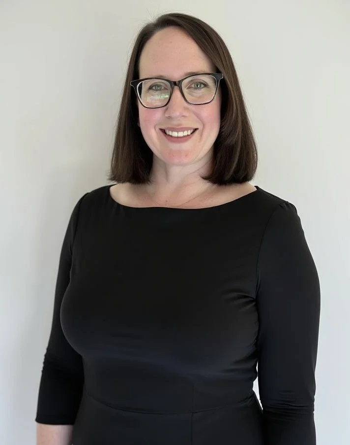 A woman with shoulder-length brown hair, glasses, and a black long-sleeve top, smiling against a plain light-colored background.