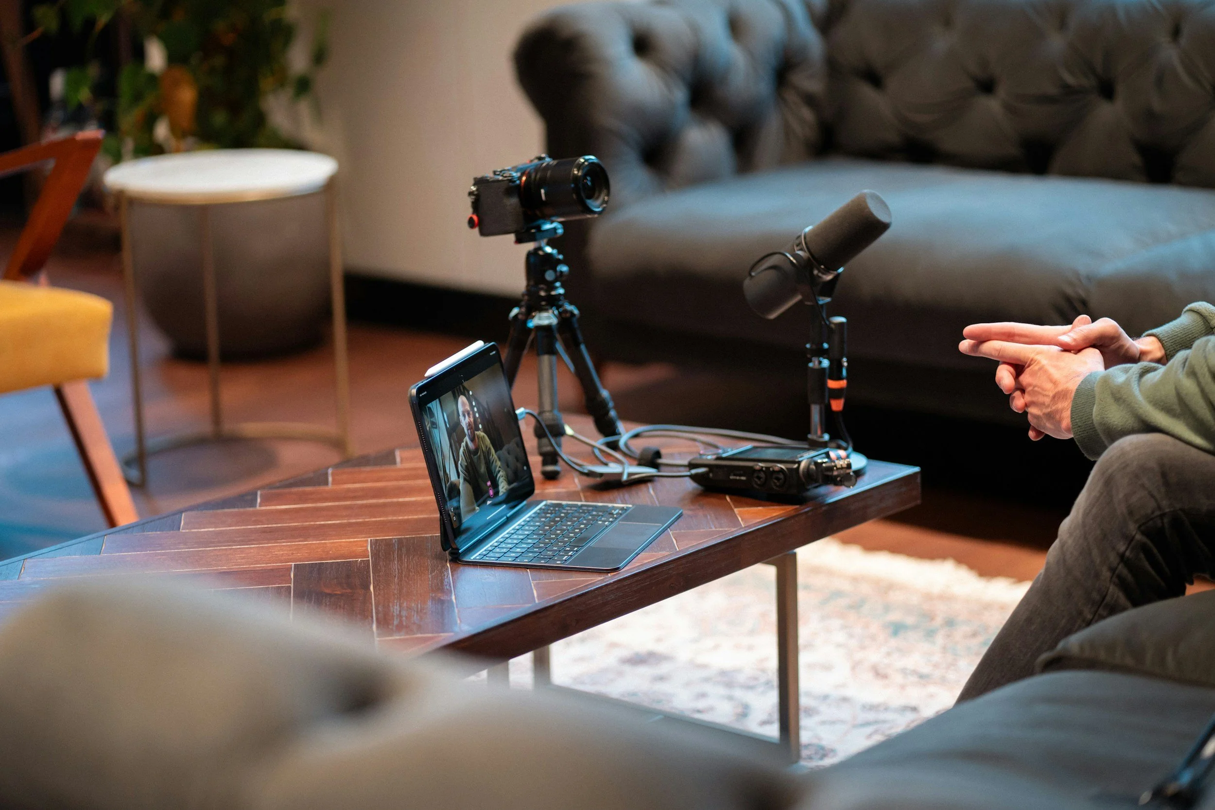 A camera mounted on a tripod and a microphone set up on a wooden table for a recording or interview session, with a person sitting nearby and a tablet showing their video feed.
