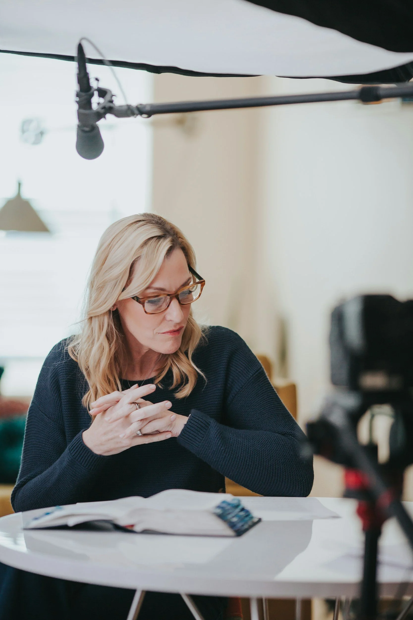 A woman with blonde hair and glasses sitting at a white table with a notebook and documents, in a room with a blurred background of lamps, wall, and other furniture.