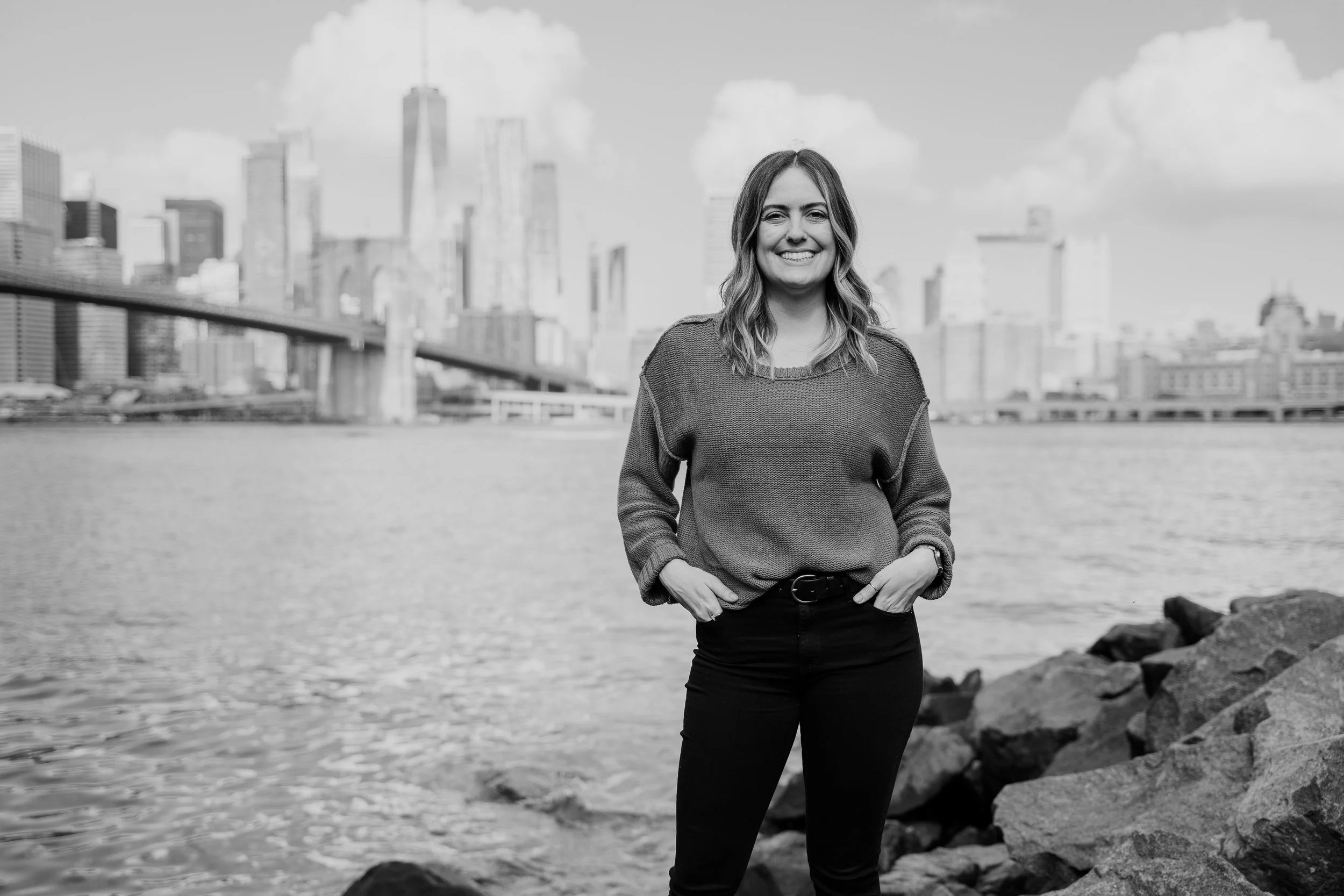 A woman standing on rocks by the water with the Brooklyn Bridge and Manhattan skyline in the background, smiling at the camera.