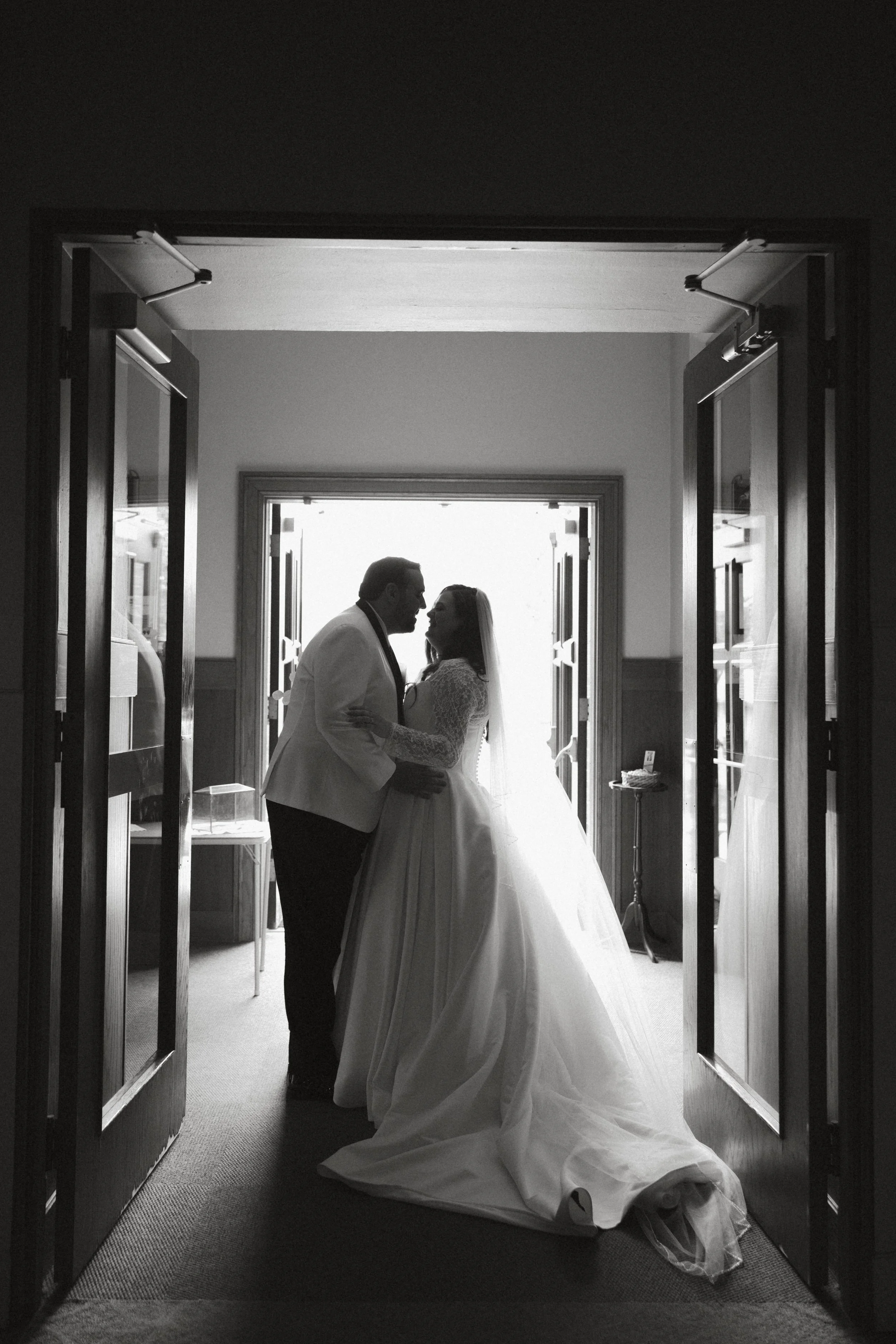 Isabel Marie Photography - Silhouette of a bride and groom sharing an intimate moment in a doorway, backlit with natural light in black and white.