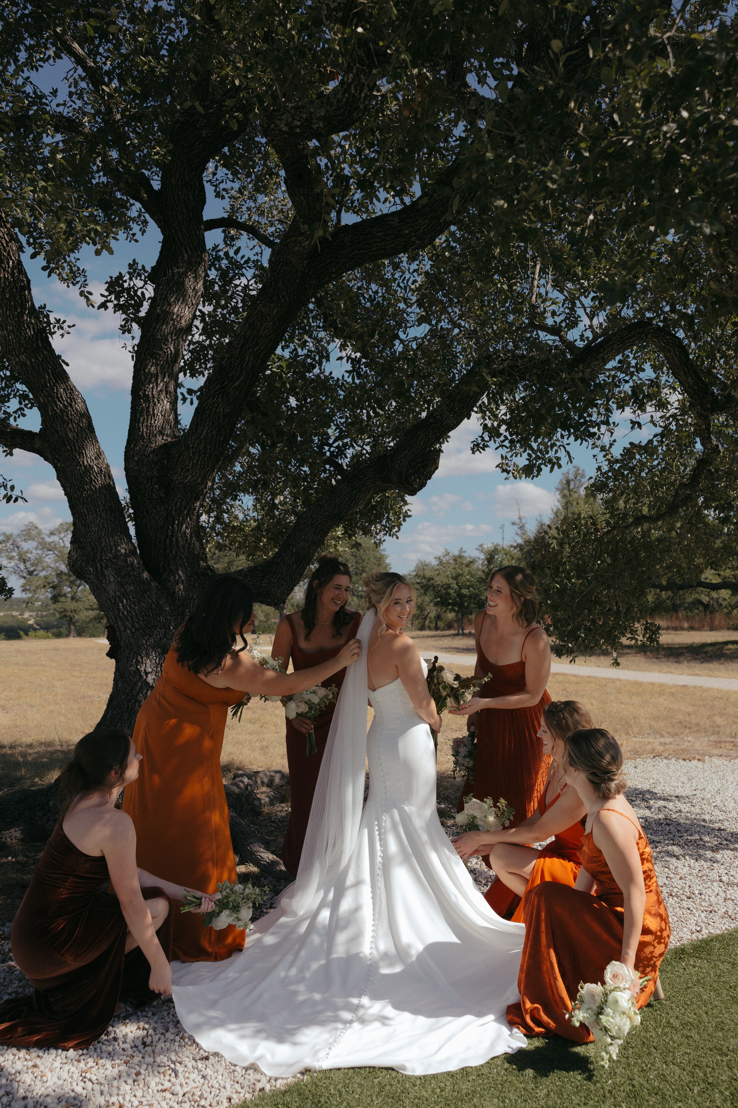 Isabel Marie Photography - Bride in white wedding gown with veil and six bridesmaids in orange and rust-colored dresses, under a large tree outdoors on a sunny day during a wedding ceremony.