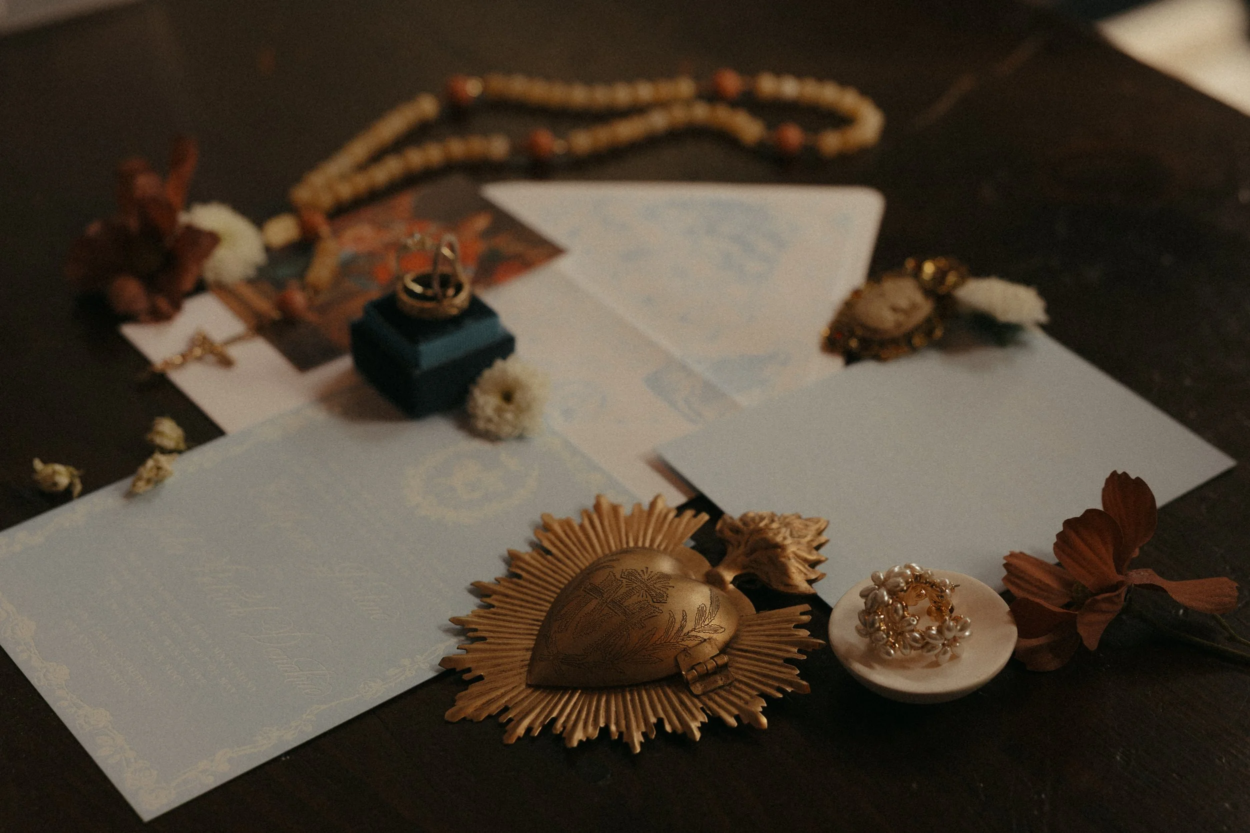 Isabel Marie Photography - Flatlays of Jewelry and paper items arranged on a dark surface, including a heart-shaped locket with a golden sunburst around it, a pearl bracelet, and assorted beads and flowers.