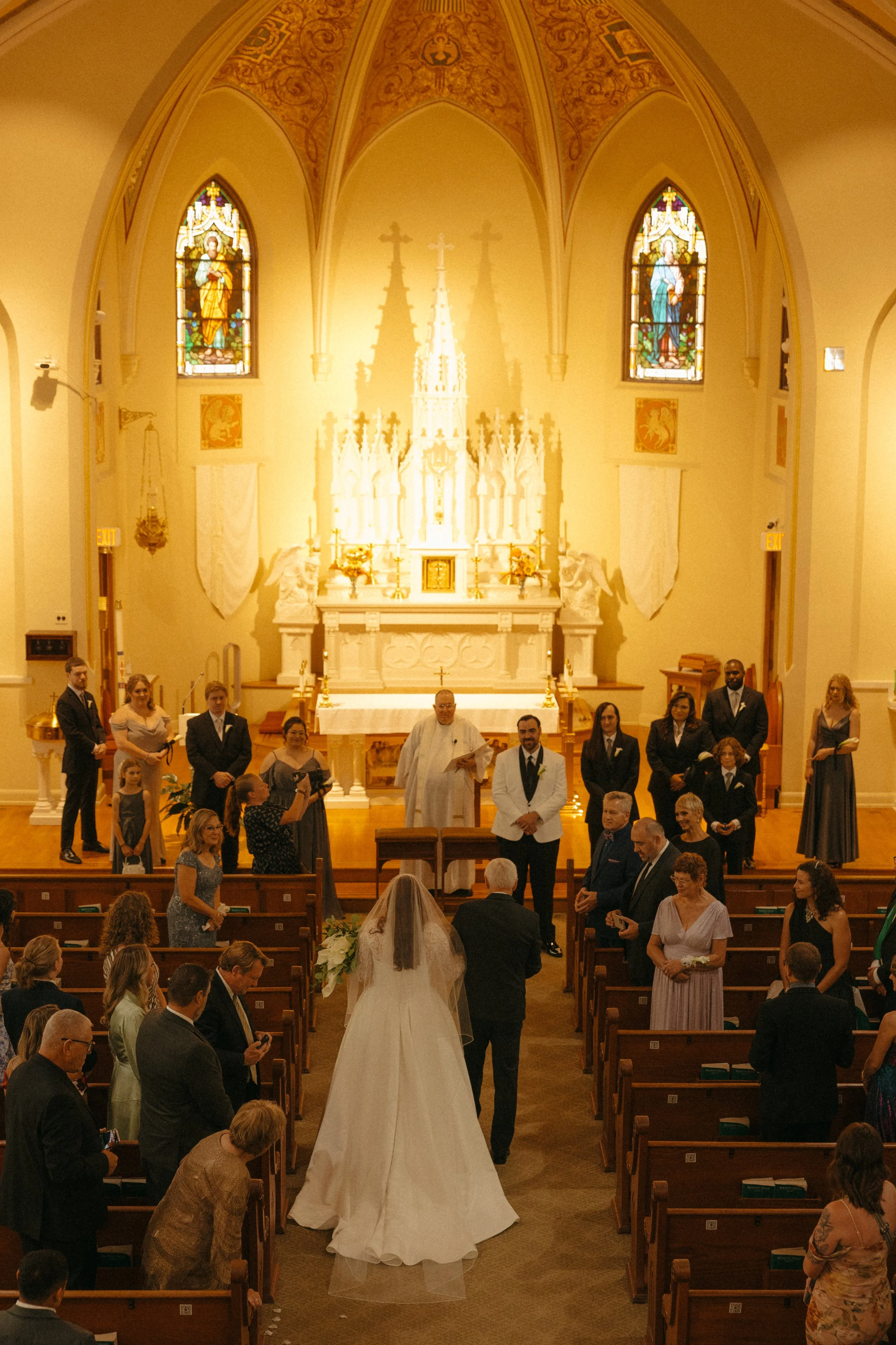Isabel Marie Photography - A bride walking down the aisle in a church during her wedding ceremony, surrounded by guests, with stained glass windows and an altar in the background.