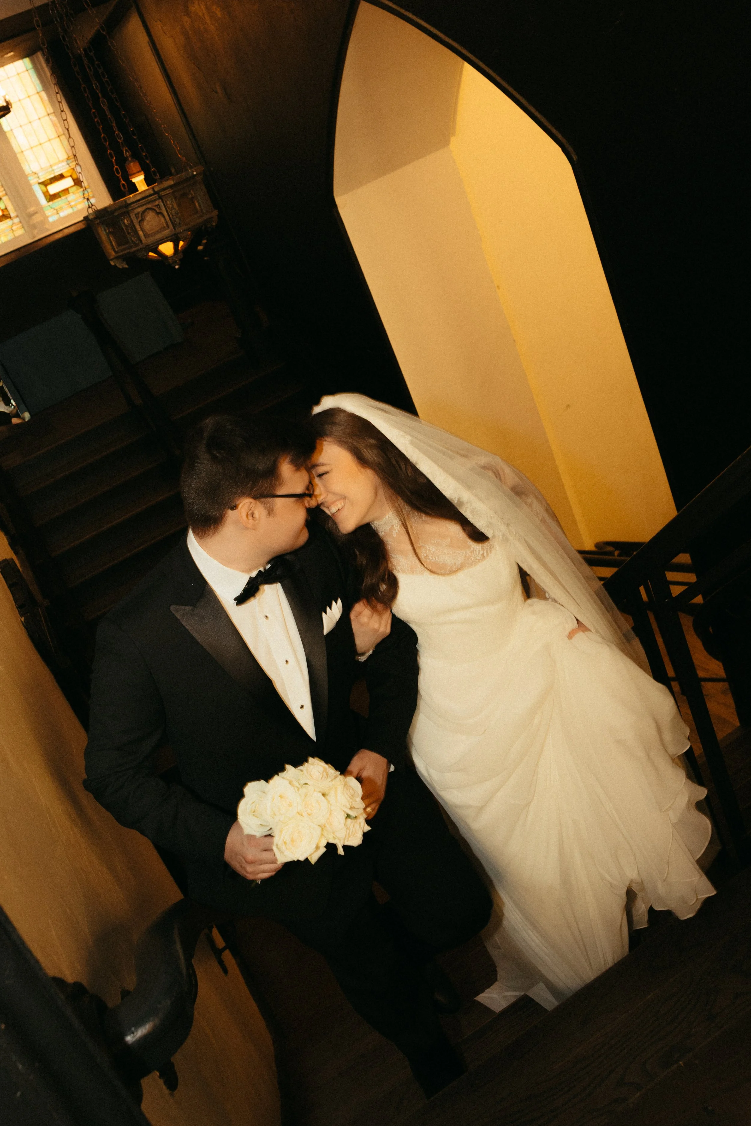 Isabel Marie Photography - A newlywed couple on a staircase. The groom is in a black tuxedo holding a bouquet of white roses, and the bride is in a white wedding dress with a veil, leaning towards each other and smiling.