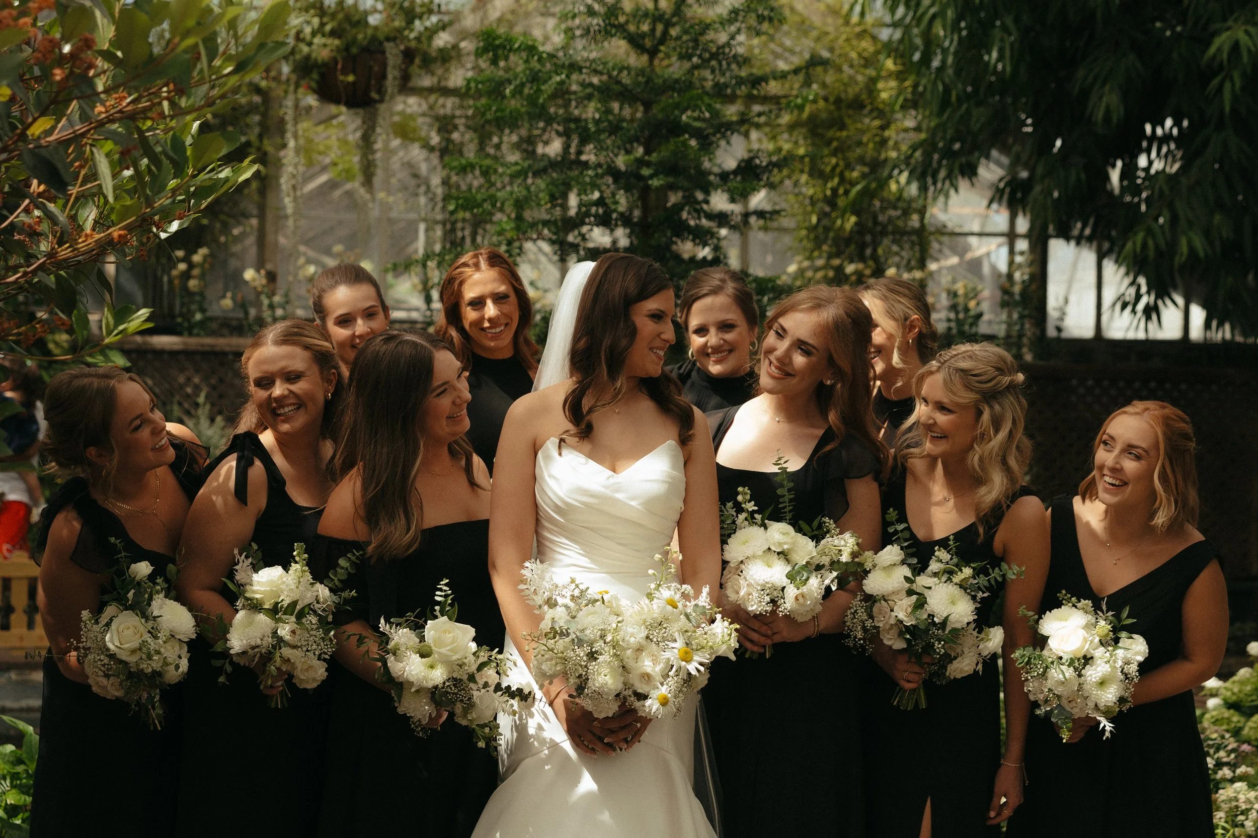 Isabel Marie Photography - A bride in a white wedding dress surrounded by her bridesmaids in black dresses, all holding bouquets of white flowers, in a lush green greenhouse